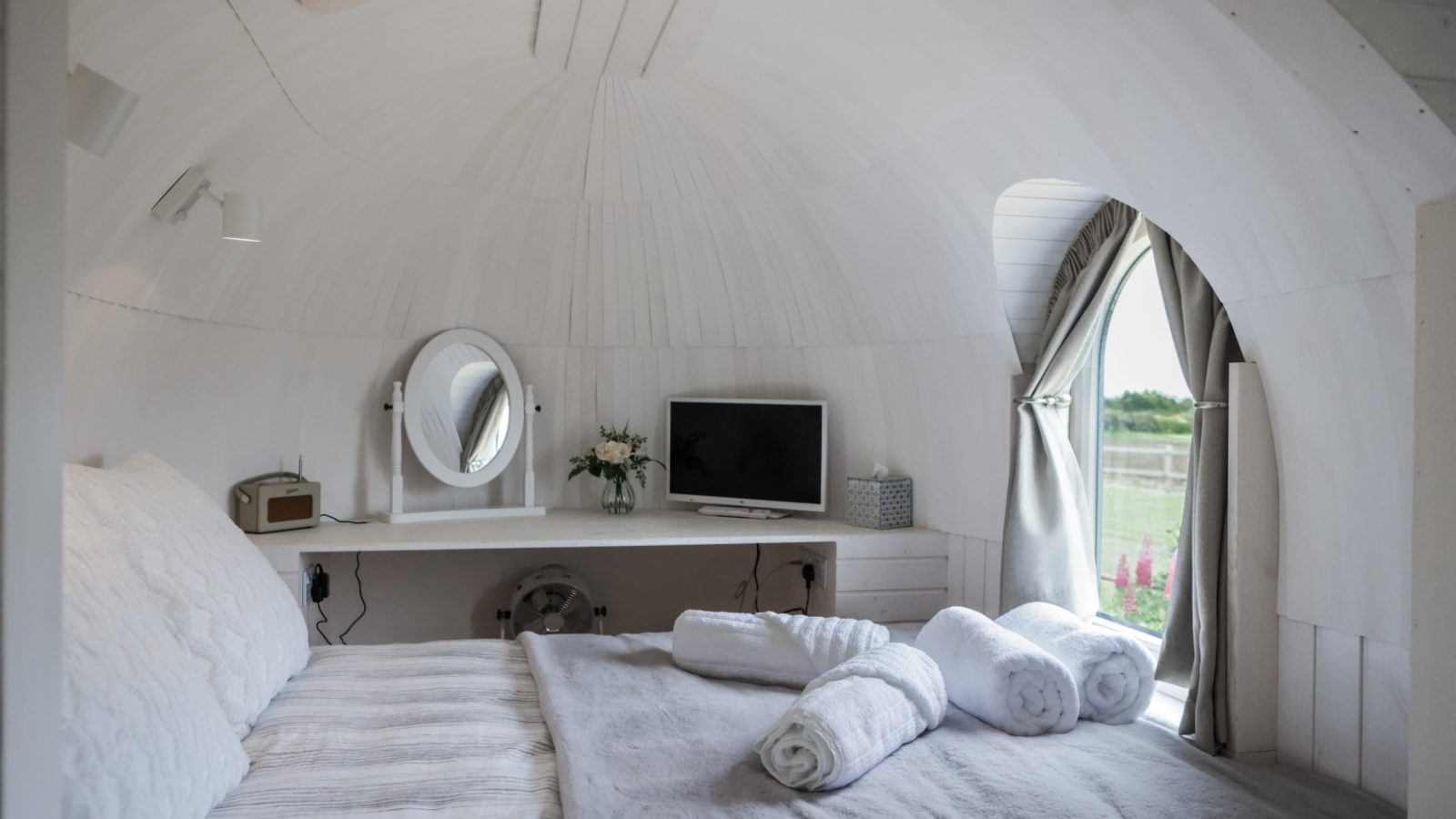 A cozy dome-shaped bedroom offers a peaceful escape, with a large bed covered in white and gray linens. The room features a small TV, a round mirror, and a vase of flowers on a shelf. Two rolled towels rest on the bed beside the window with curtains that reveal views resembling those from a castle.