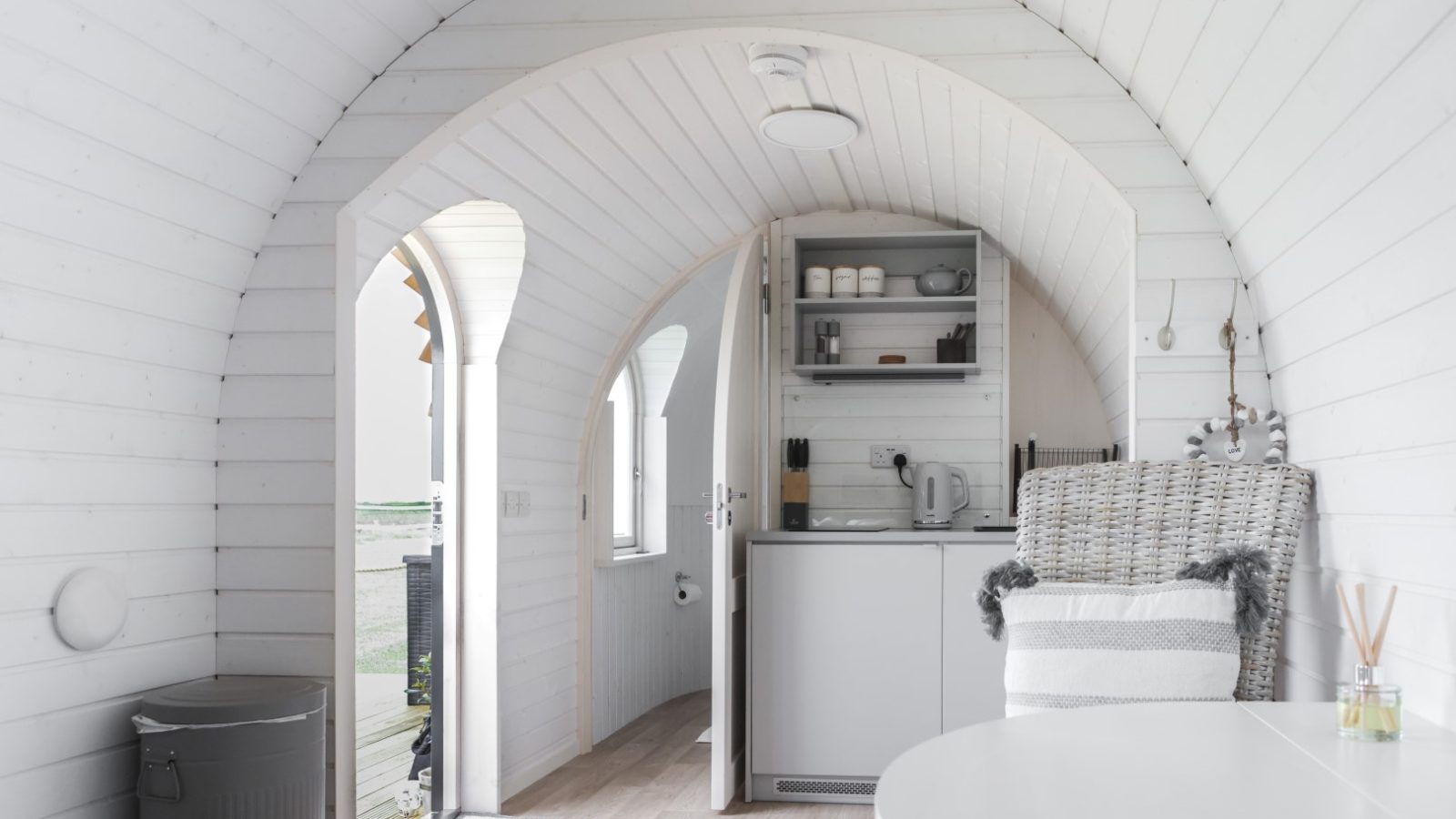 Inside this small, modern wooden cabin with white arched walls, experience a castle escape vibe. It features a compact kitchenette with open shelving, a cozy chair draped in a throw blanket, and light wood flooring. A door opens to the outside, inviting in natural light.