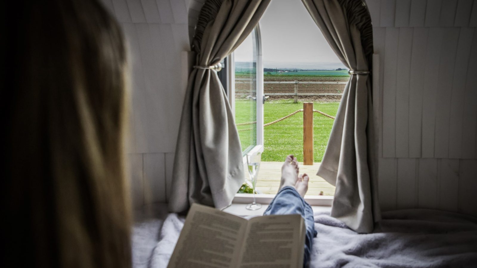 A person is reclining on a bed, reading a book near a window of their tranquil escape. The open curtains reveal a view of lush green landscape and an overcast sky, capturing the serene atmosphere. A glass of white wine rests beside them, enhancing the peace of this castle-like retreat.