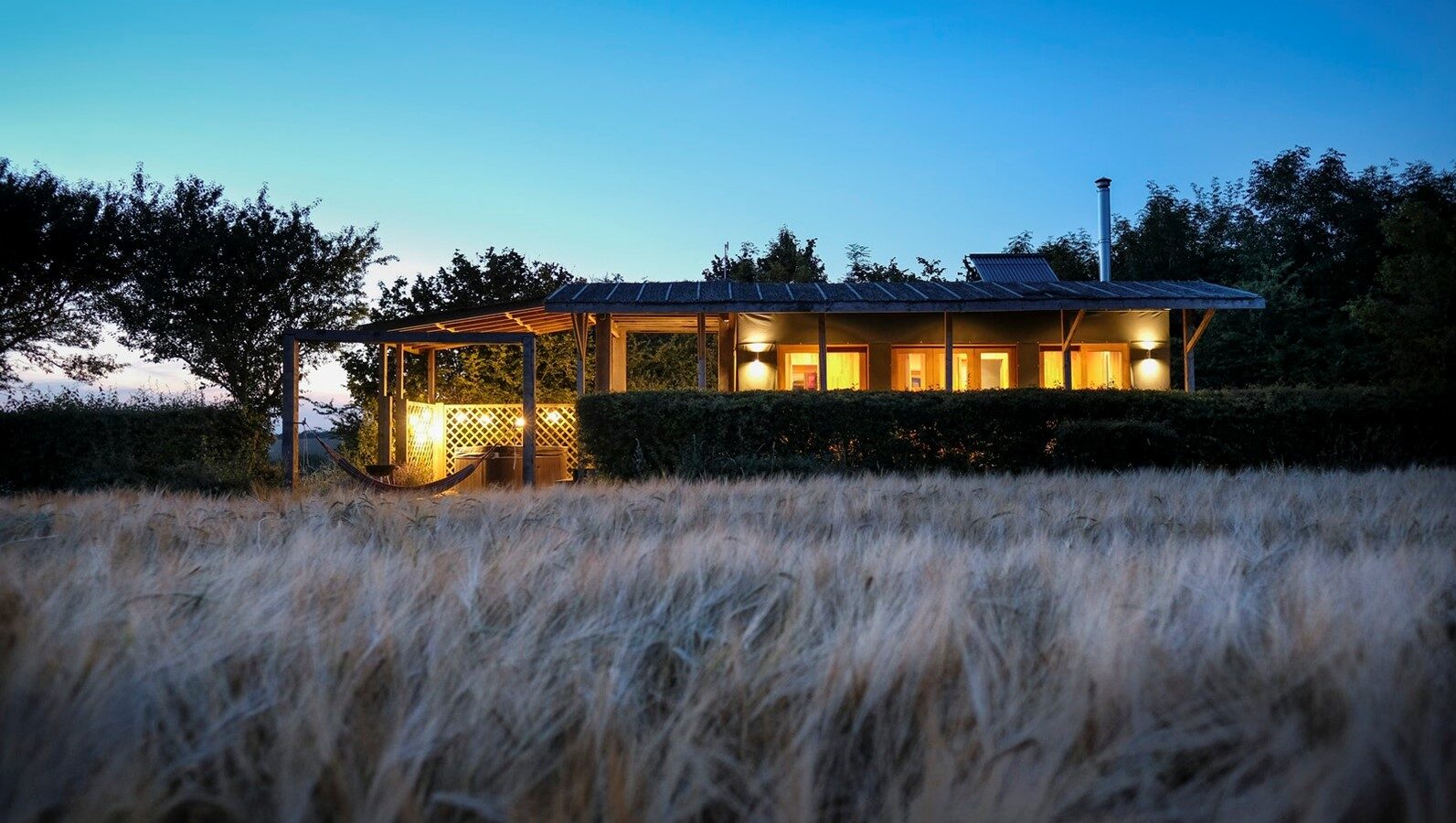 A cozy, illuminated lodge at dusk, nestled in the Chilterns and surrounded by trees and a field of tall grass under a clear sky.