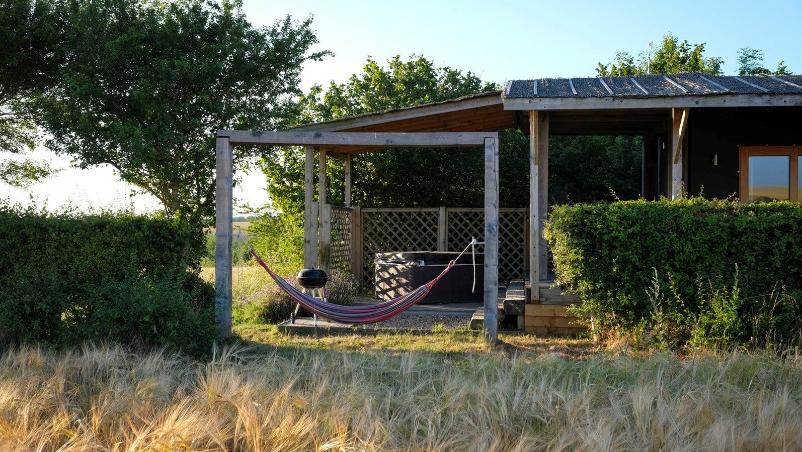 A colorful hammock hangs between two posts in front of a wooden deck and patio, nestled within the serene beauty of Chilterns View Lodges. Surrounded by trees and grass under a clear sky, it offers a perfect retreat in the heart of the Chilterns.