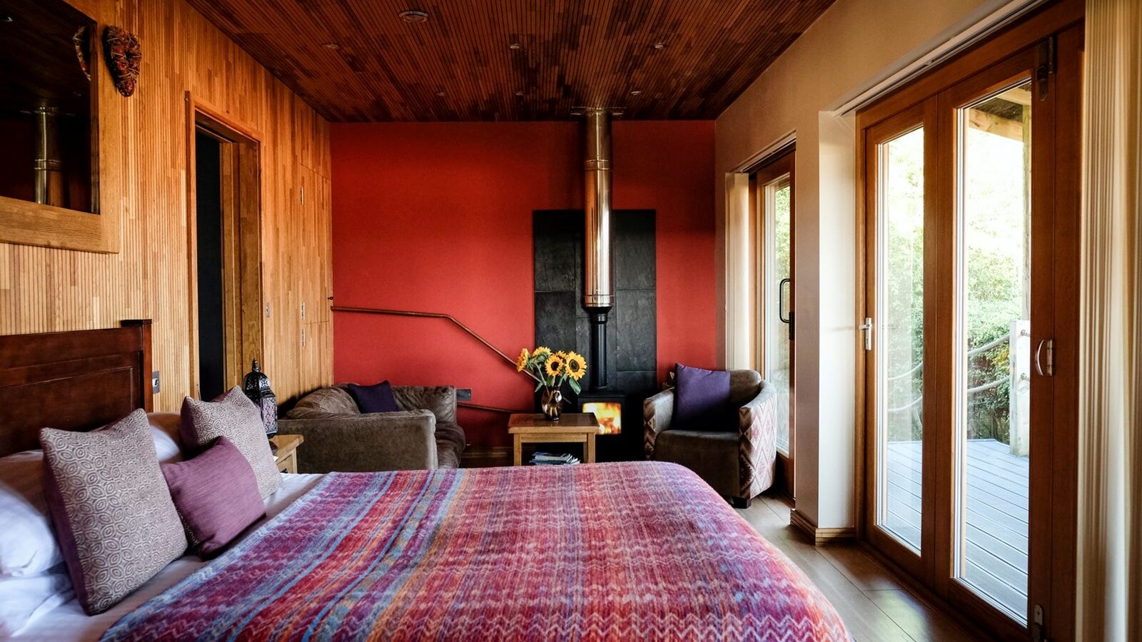 Experience a cozy bedroom at Chilterns View Lodges, featuring a red accent wall, fireplace, and large windows. The space includes a comfortable bed, chairs, and vibrant sunflowers on the table for an inviting atmosphere.