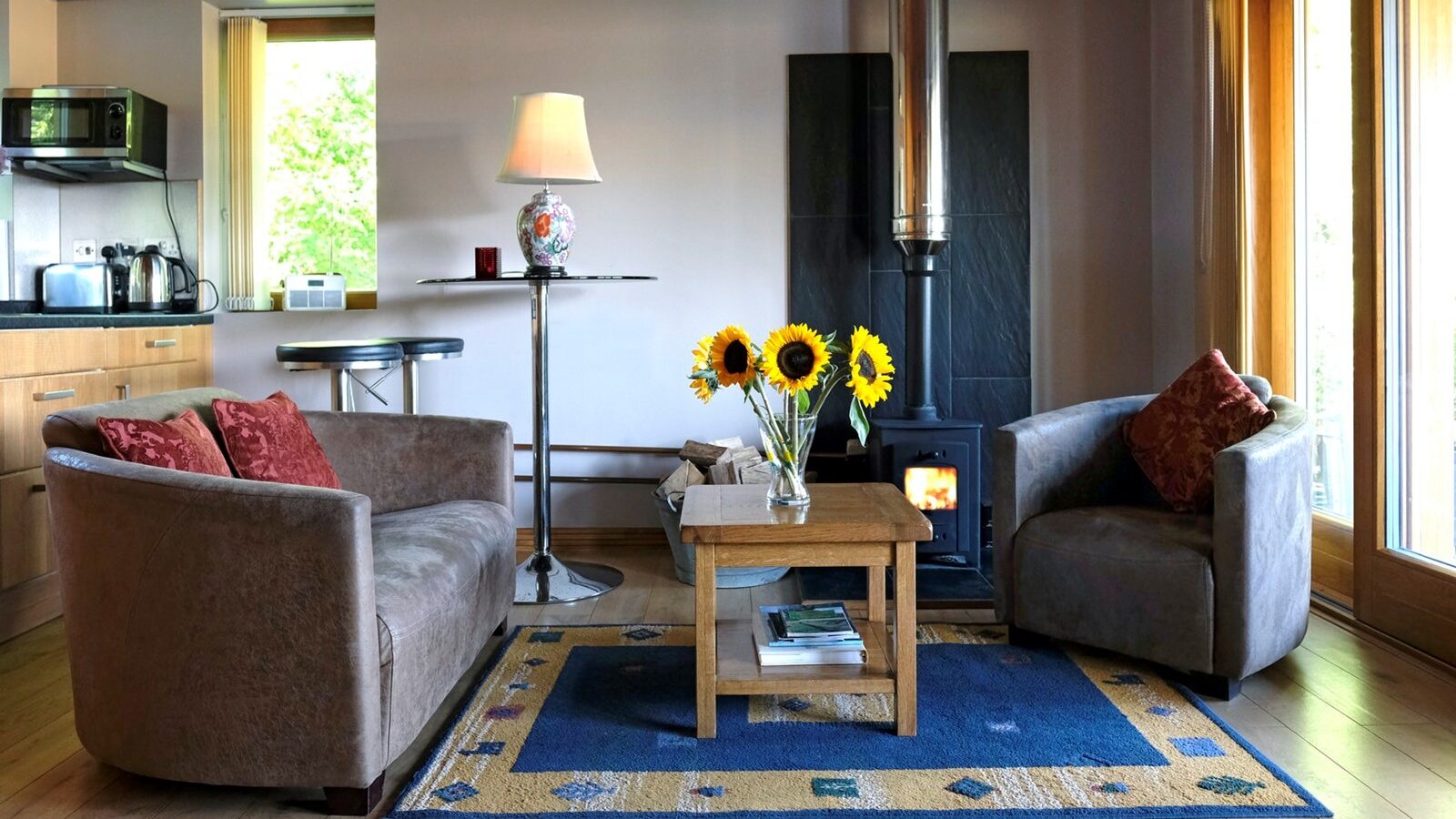 Cozy living room with two sofas, a coffee table adorned with sunflowers, and a fireplace on a plush blue rug. In the background, the kitchen offers a glimpse of warmth and hospitality, reminiscent of the welcoming charm found at Chilterns View Lodges.