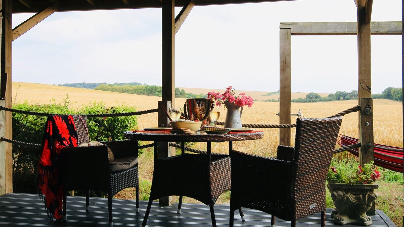 Outdoor patio at View Lodges with a table set for two, surrounded by wicker chairs, overlooking the rolling wheat fields of the Chilterns and a hammock gently swaying on the side.