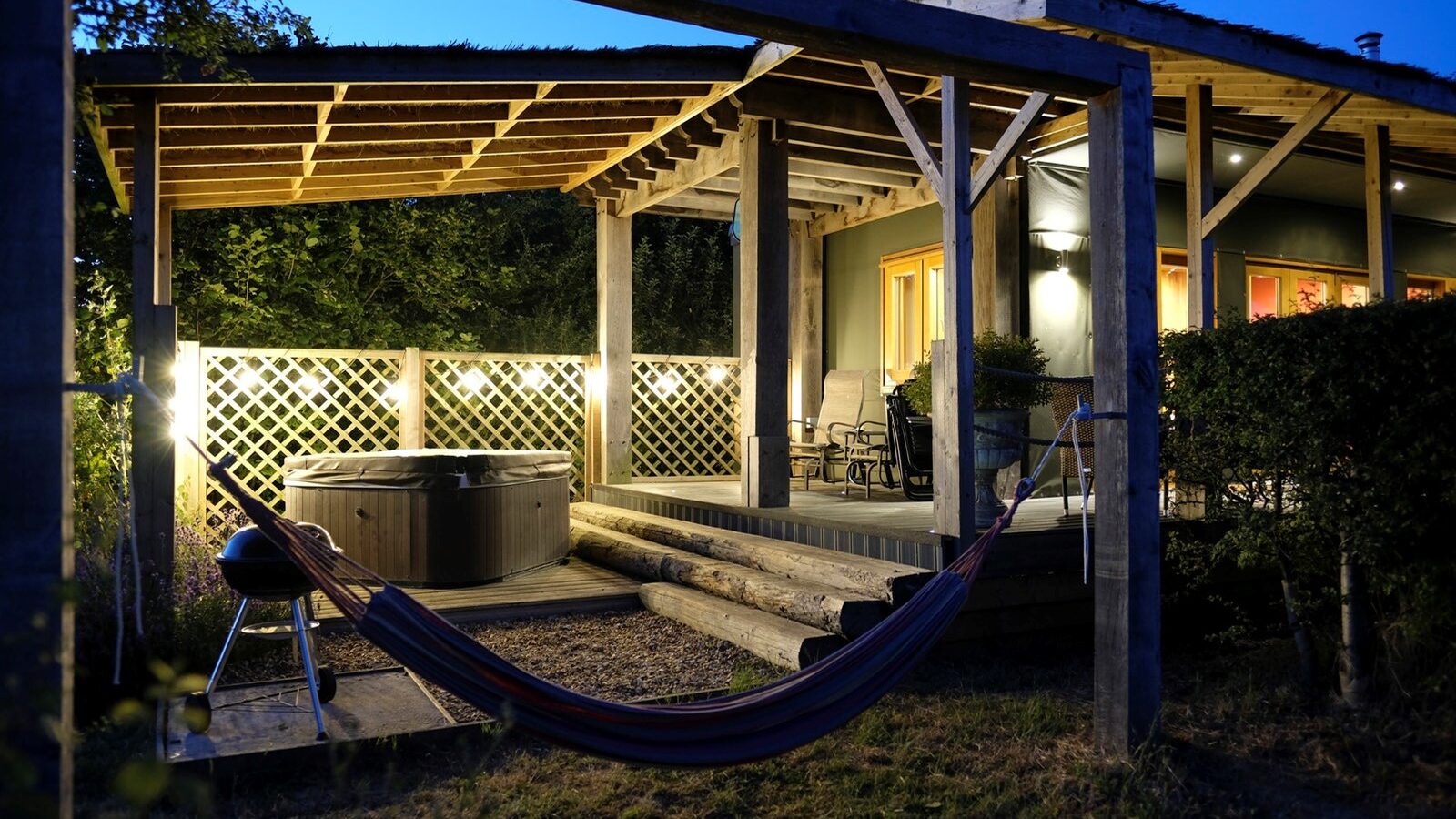 A backyard patio at dusk with a hot tub, hammock, and seating area under a wooden pergola, lit by outdoor lights, mirrors the relaxing vibe of Chilterns View Lodges.