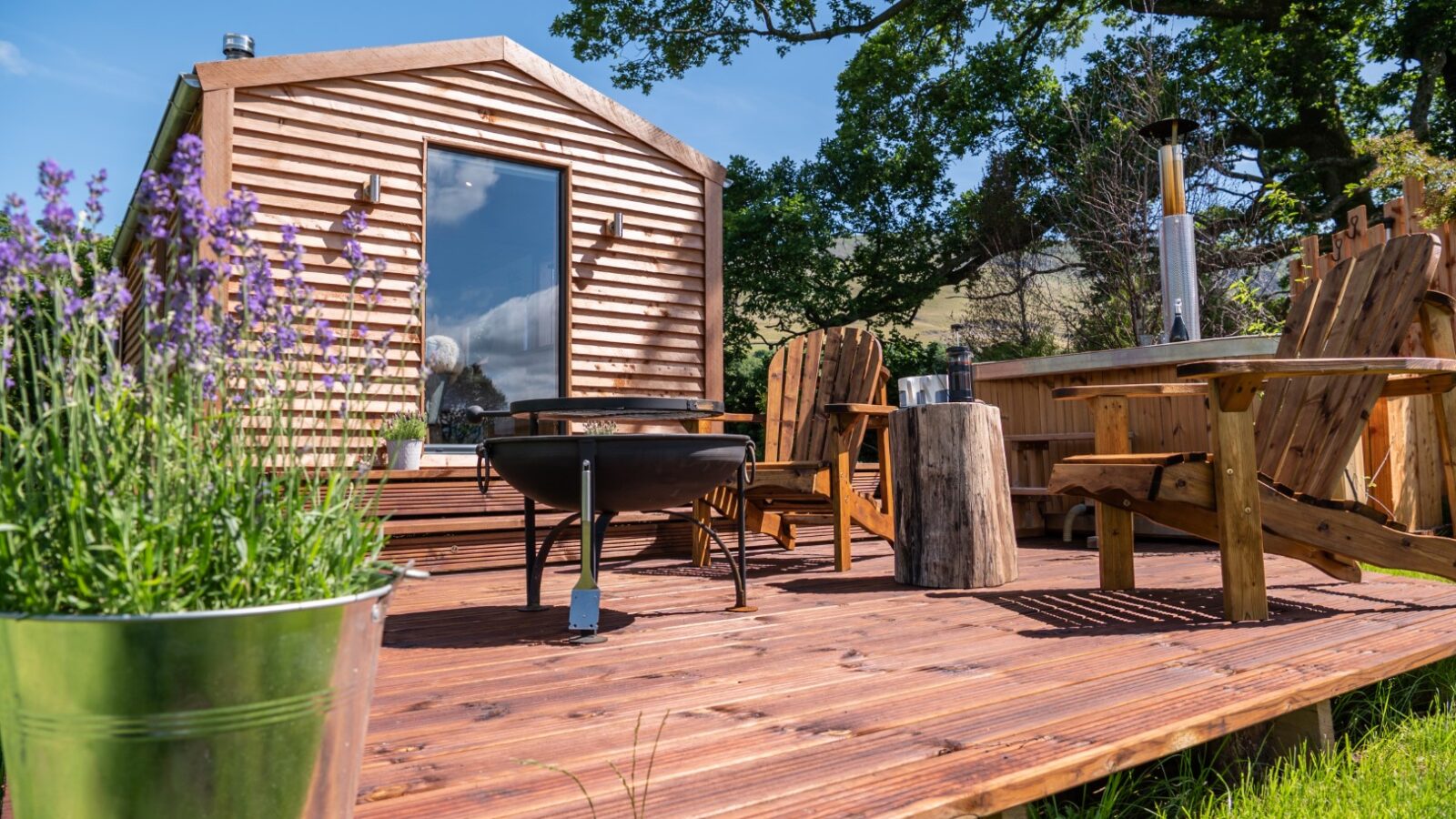 Nestled in The Vale, this wooden cabin offers a cozy patio with a fire pit, inviting chairs, and a charming tree stump table. Lavenders bloom beautifully in a metal pot nearby, adding to the serene ambiance.