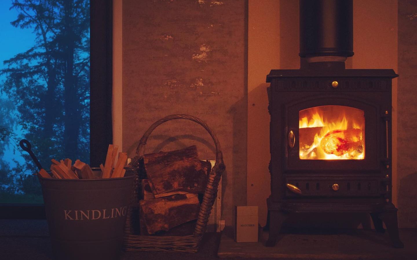 A cozy wood-burning stove is lit, nestled in a charming treehouse. Surrounded by a basket of firewood and a bucket labeled 