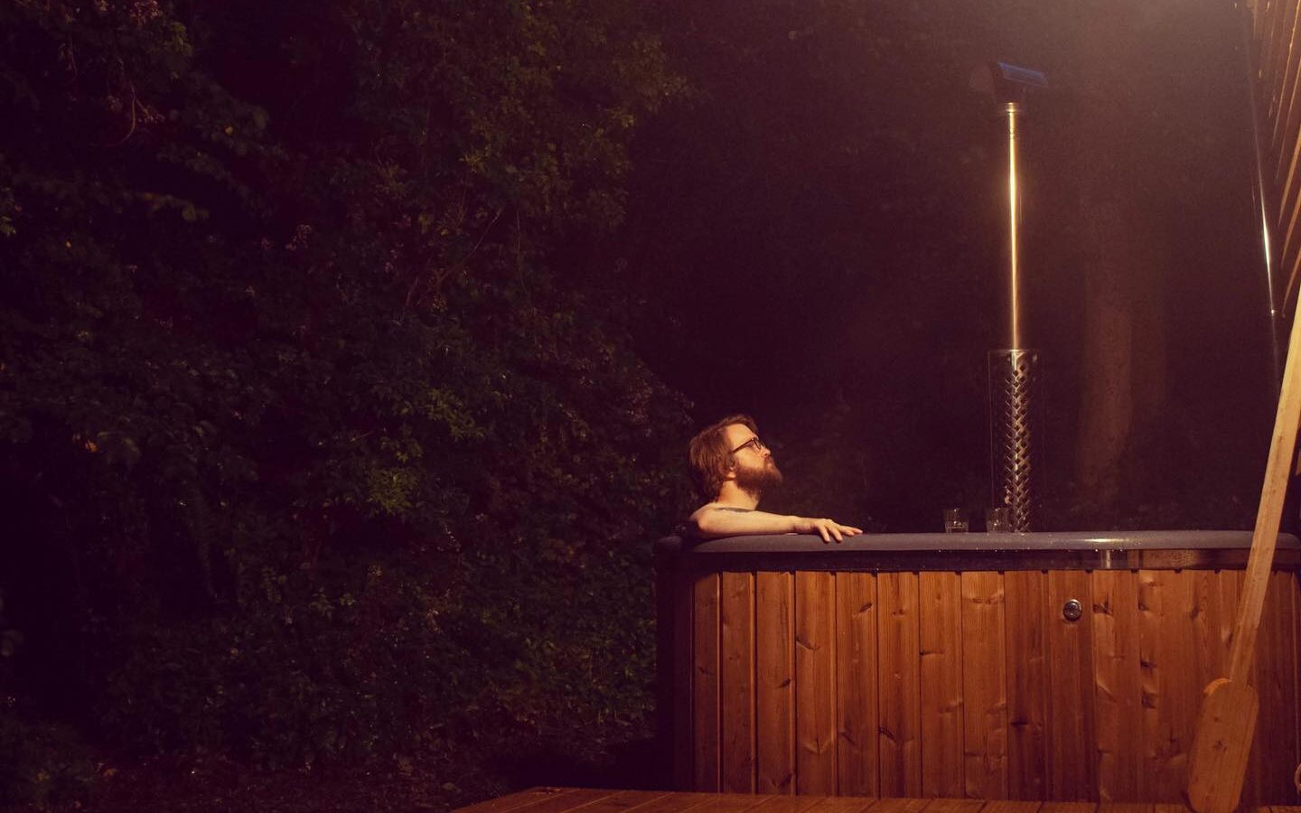 Person relaxing in an outdoor hot tub at night, nestled near a treehouse under the starlit sky, with a drink on the edge of the tub.