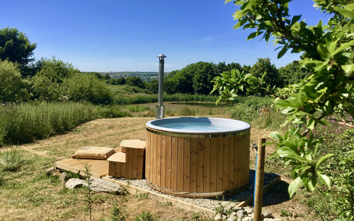 Nestled in a grassy area at Sloeberry Farm, this wooden hot tub offers a tranquil escape with a distant view of rolling hills under a clear blue sky, perfectly complementing the charming nearby lodges.