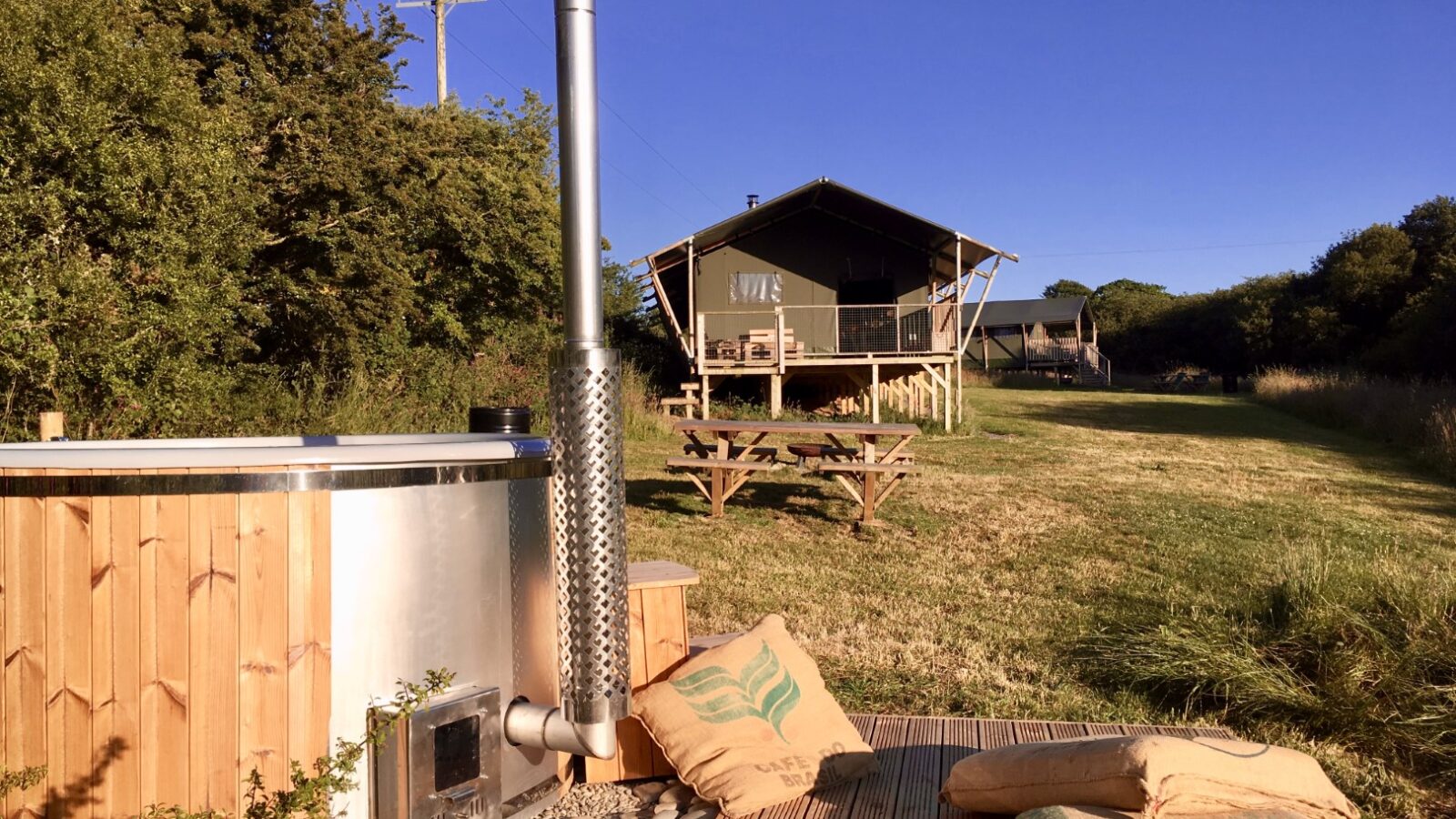 Nestled in a grassy field under a clear blue sky, Sloeberry Farm's wooden cabin offers relaxation with its hot tub and cozy deck. Enjoy the picnic table and cushions, creating the perfect countryside retreat at one of the most charming lodges around.