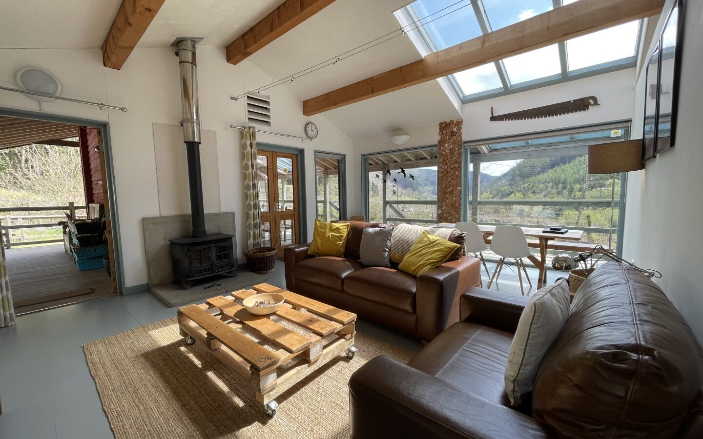 A cozy living room in Trallwm Forest Cottages features leather sofas, a wooden coffee table, a fireplace, and large windows offering a breathtaking mountain view.