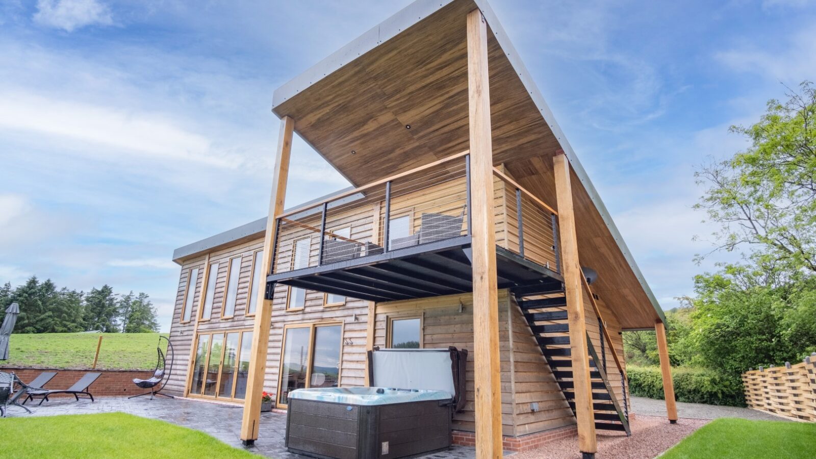 Experience the charm of Lundy Lodges, featuring a modern two-story wooden house. This exquisite accommodation boasts a large slanted roof, balcony, staircase, hot tub, and is enveloped by lush greenery—perfect for relaxing in style.