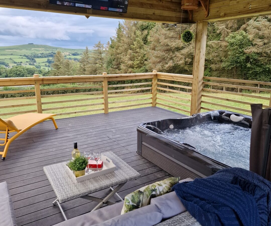 On the deck of our charming Lundy lodge, you'll find an inviting outdoor setup complete with a hot tub, lounge seating, coffee table, and TV—all overlooking a scenic landscape of rolling hills and lush trees.