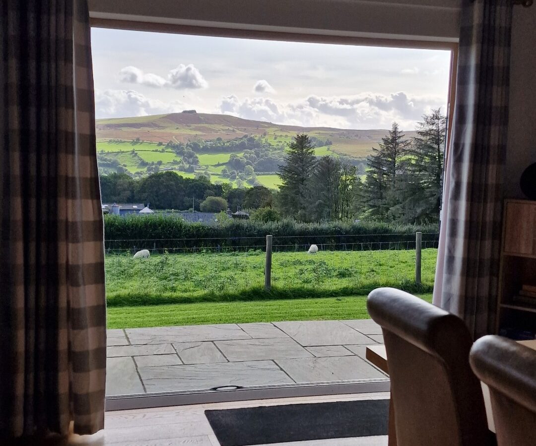 A view of a lush green landscape with hills and trees unfurls through a large window adorned with plaid curtains, reminiscent of the scenic beauty surrounding the cozy lodges on Lundy Island.