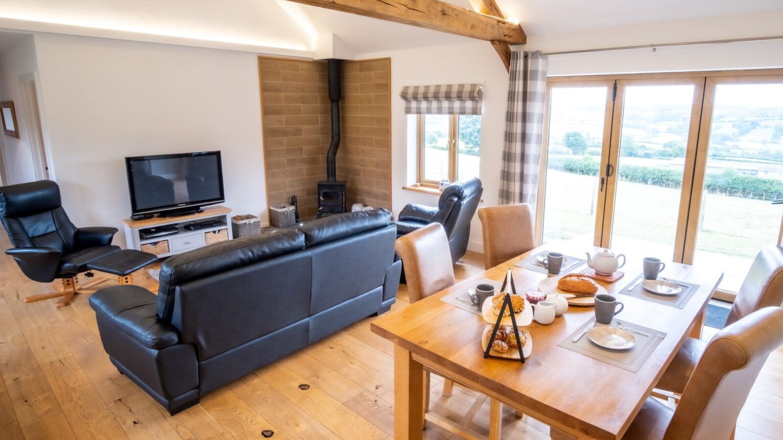 Cozy living room in one of the charming Lundy lodges, featuring a dining table, leather sofas, a TV, wood-burning stove, and large windows with countryside views.