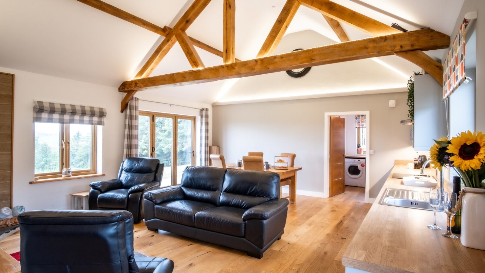 Experience the charm of Lundy Lodges with a spacious living room featuring leather sofas, exposed beams, a dining table, and large windows. An open doorway invites you to explore more of this delightful lodging.