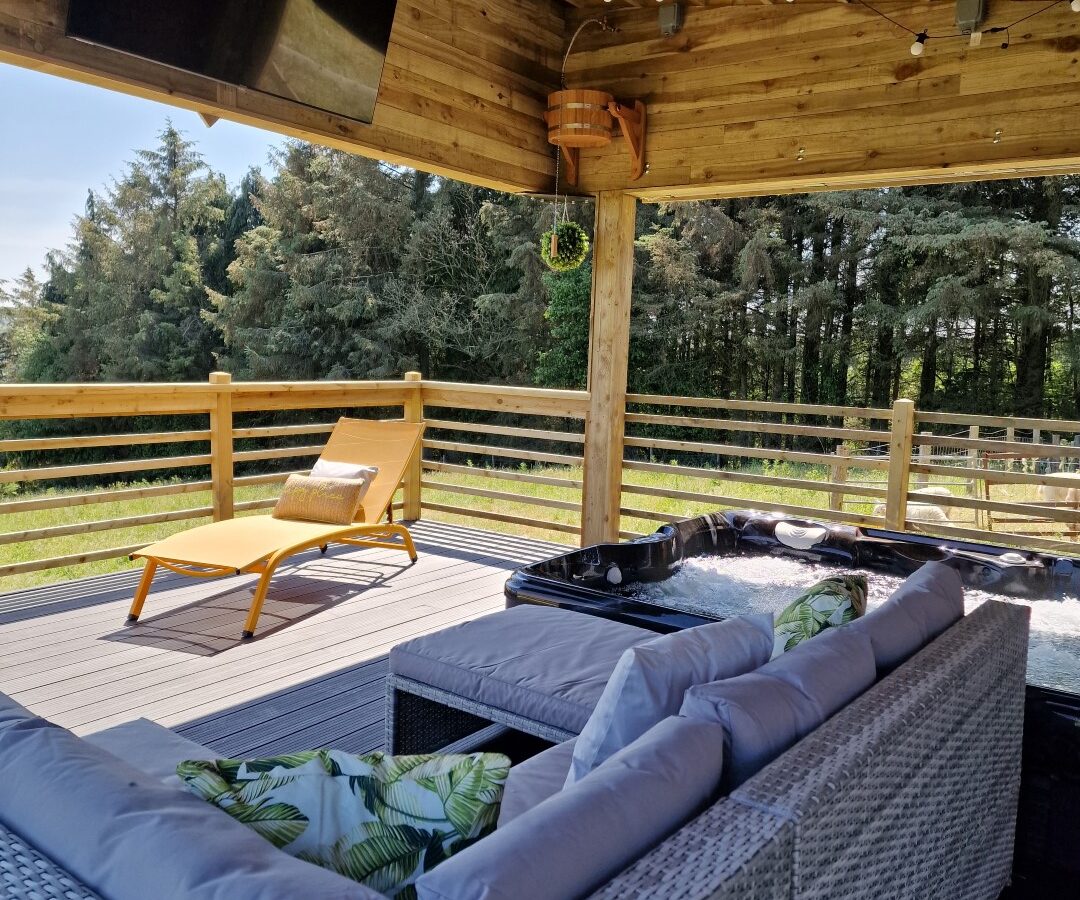 Nestled in tranquility, this outdoor lounge at Lundy features a wicker sofa set, a reclining chair, a TV, and a hot tub under a wooden pergola with breathtaking views of the forest. This serene retreat offers the perfect mix of comfort and nature reminiscent of secluded lodges.