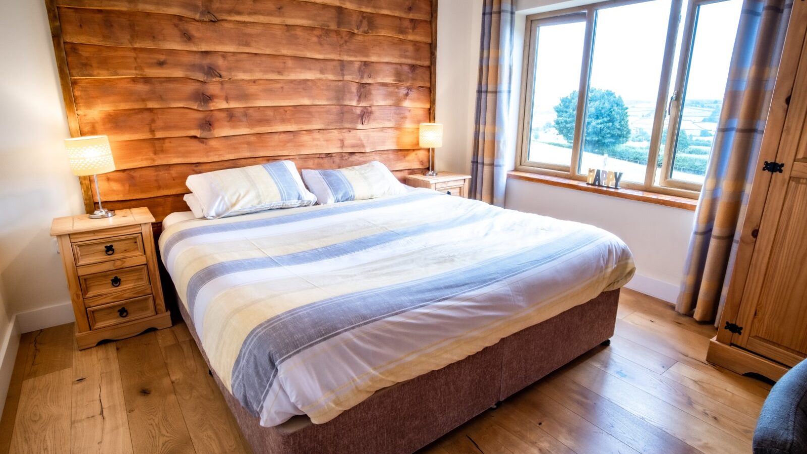 A cozy Lundy bedroom features a large bed with striped bedding, two bedside tables with lamps, and a window offering a countryside view, perfect for lodge-style accommodation.