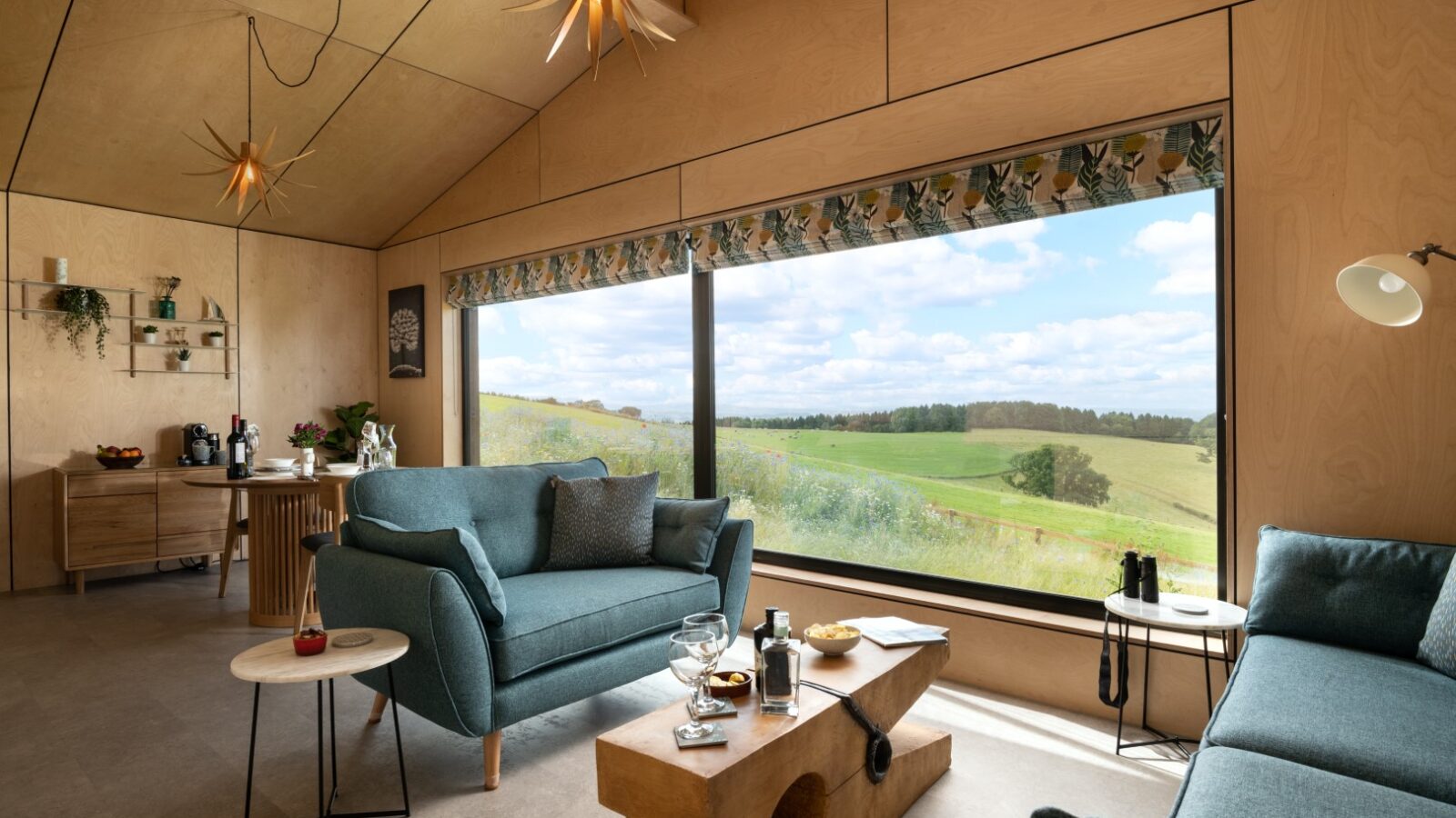 Discover the charm of a modern living room at Bettws Court Retreats, complete with teal sofas, wooden furniture, and artistic lighting. Enjoy stunning views through large windows that overlook the serene rolling hills.