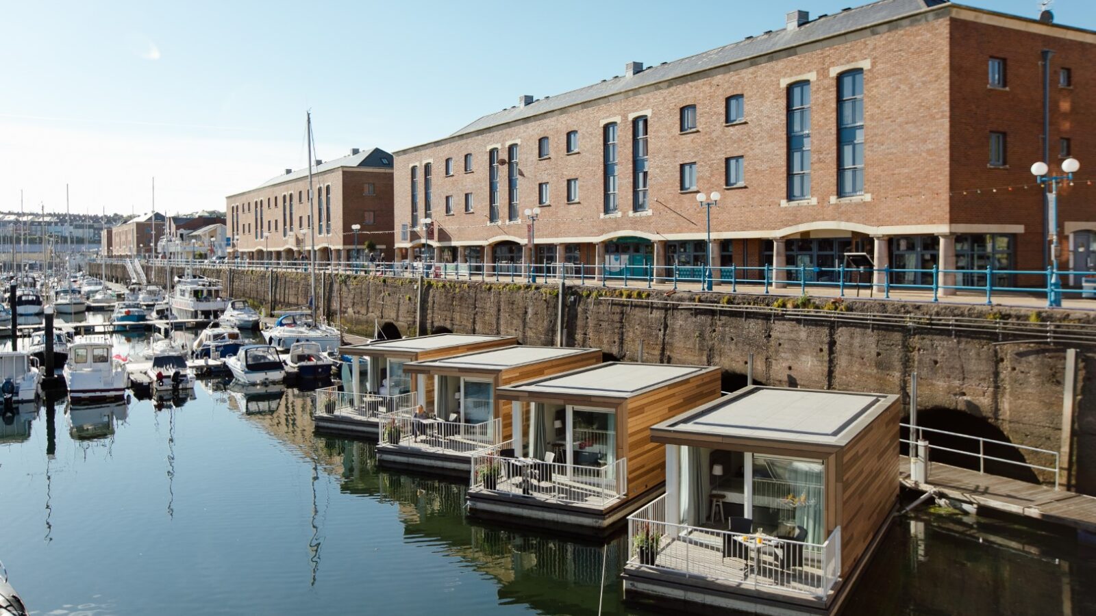 Floating houses and docked boats in the marina, reminiscent of Ty Milford Floatels, with brick buildings in the background under a clear blue sky.