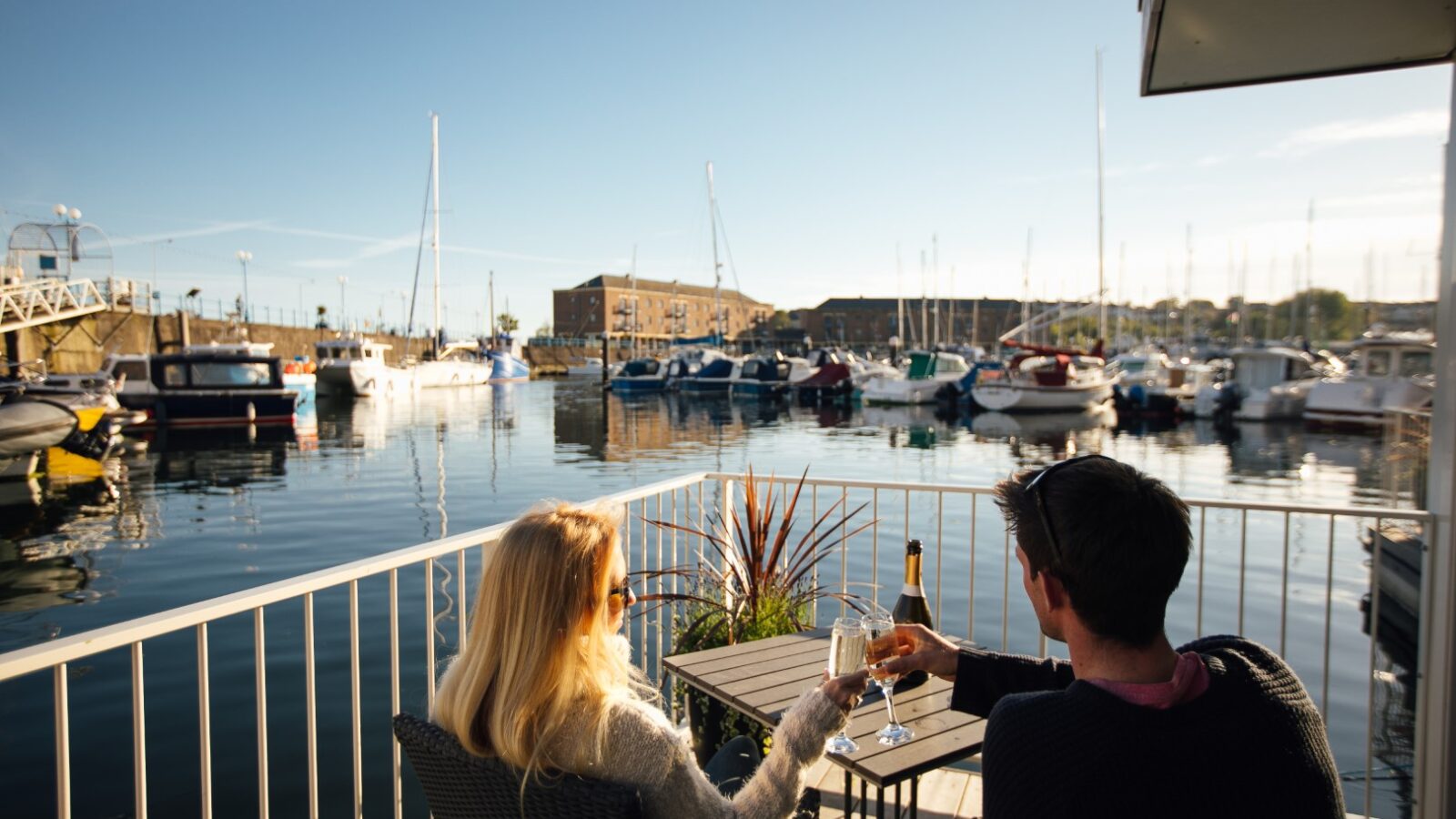 Two people sit on a balcony at the Ty Milford Floatels, clinking wine glasses and enjoying a stunning view of boats bobbing in the marina under a clear blue sky.