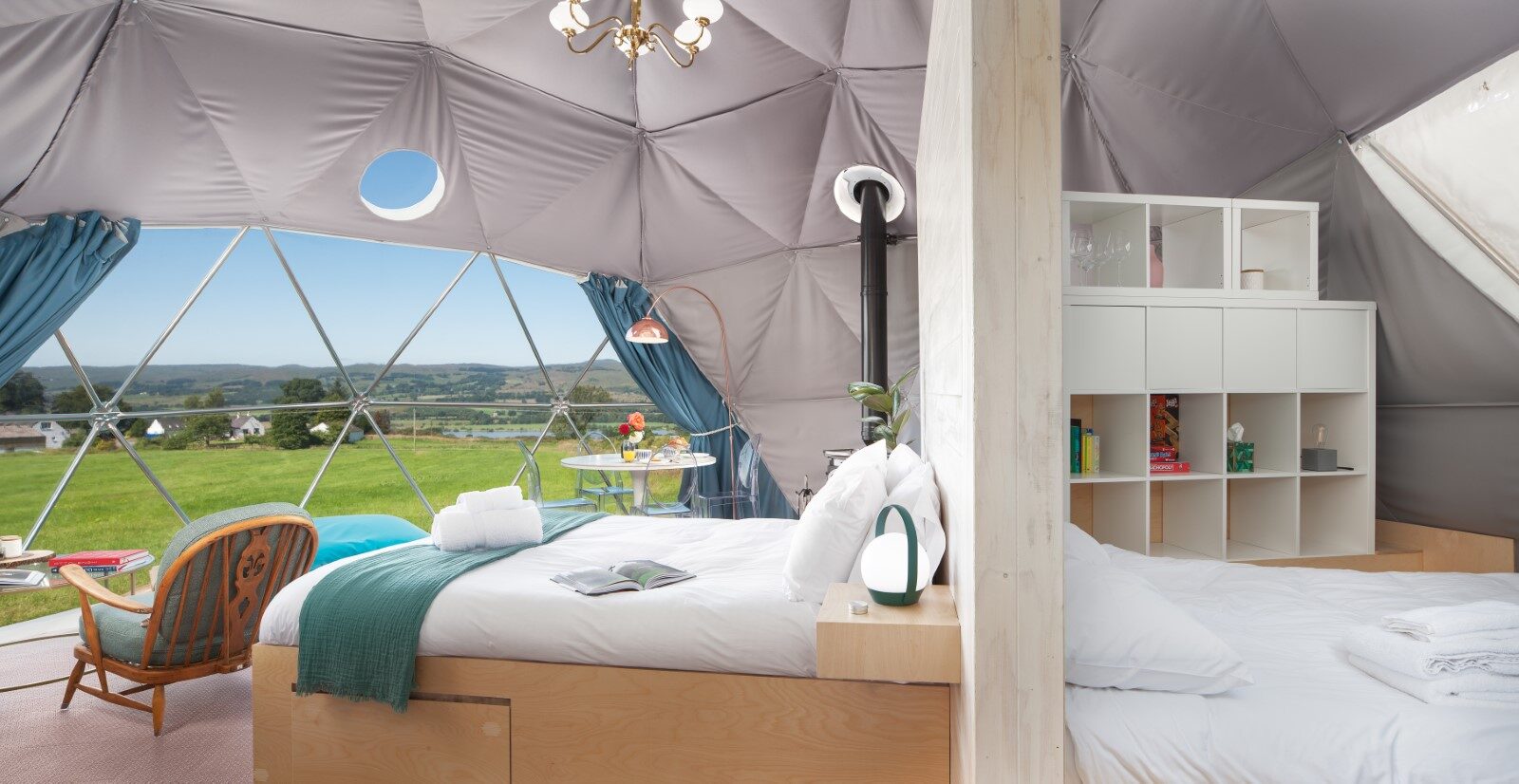 The interior of the Mid Auchengowan Dome features two cozy beds, inviting chairs, and a stunning view of the grassy landscape framed by a large window.