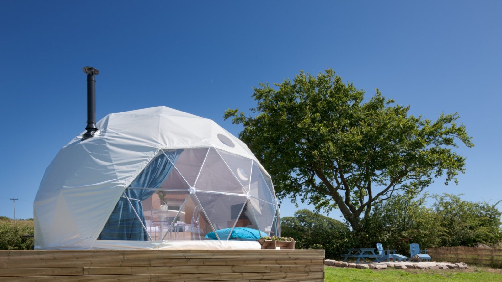 The Mid Auchengowan Dome, gracefully perched on a wooden platform, features a transparent front. Nestled under a clear blue sky next to a solitary tree in the grassy area, this dome offers an idyllic retreat.