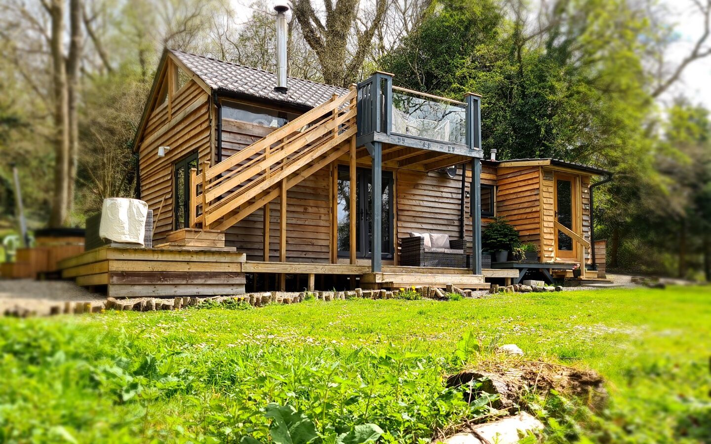 Nestled among the trees, this small wooden cabin feels like a serene treehouse. It features an upper deck with a glass railing, perfect for star gazing and enjoying the tranquil view of the grassy area below.