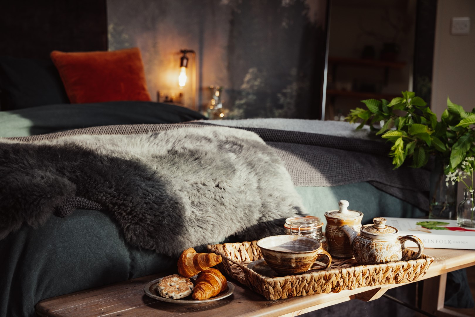 Cosy bedroom featuring a bed with a fur throw and a wooden tray holding tea, croissants, and cookies. Lush plants and books add charm to the background. Perfect for a romantic adults only escape for two