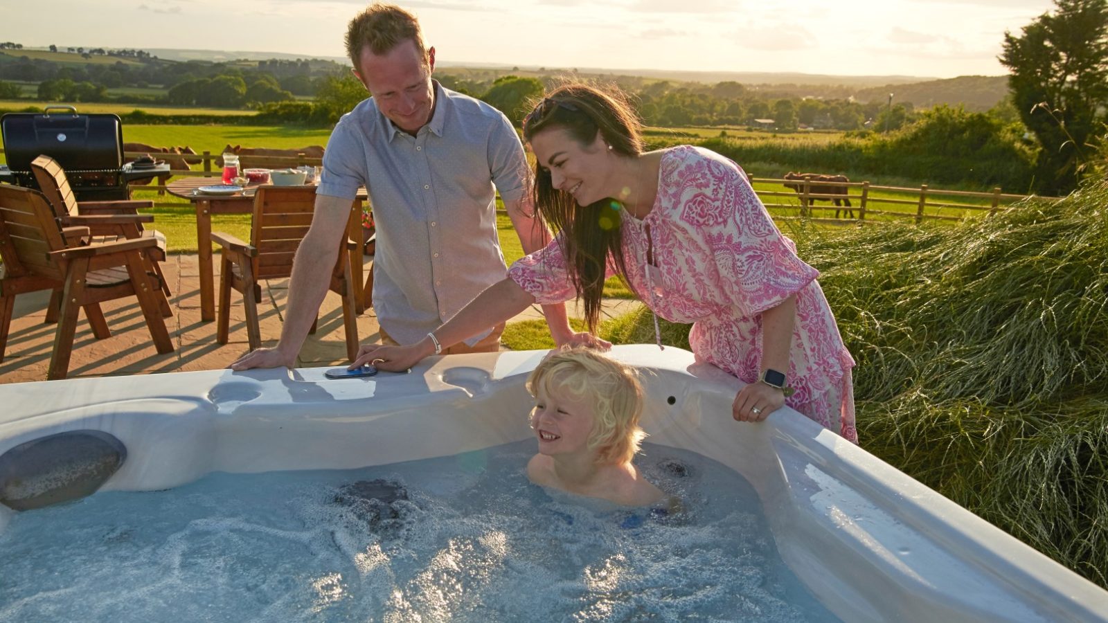 A child sits in a hot tub while two adults stand nearby, gazing fondly at the young bather. The outdoor setting boasts the rolling hills of Yorkshire in the background, adding to the picturesque scene.