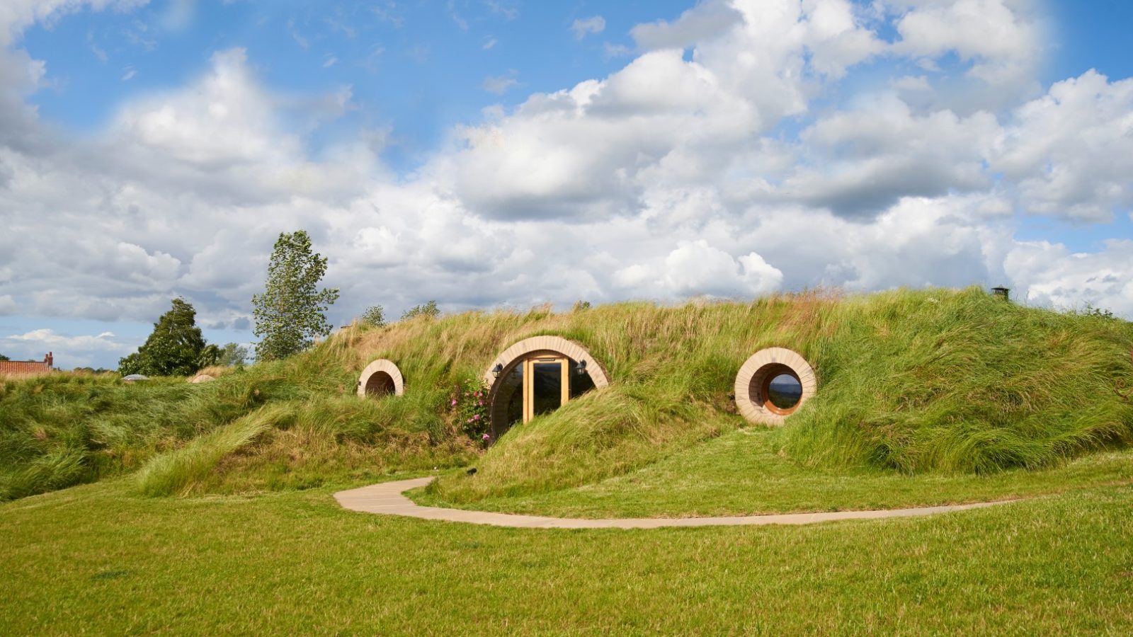 A small, grass-covered building resembling a hobbit burrow with round doors and windows sits serenely under a cloudy blue Yorkshire sky.