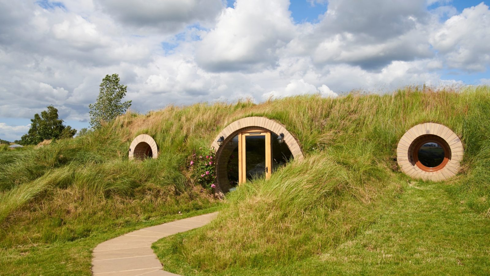 A grass-covered, hobbit-style house reminiscent of the charming Yorkshire Burrows, with round windows and a wooden door, stands gracefully under a cloudy sky.