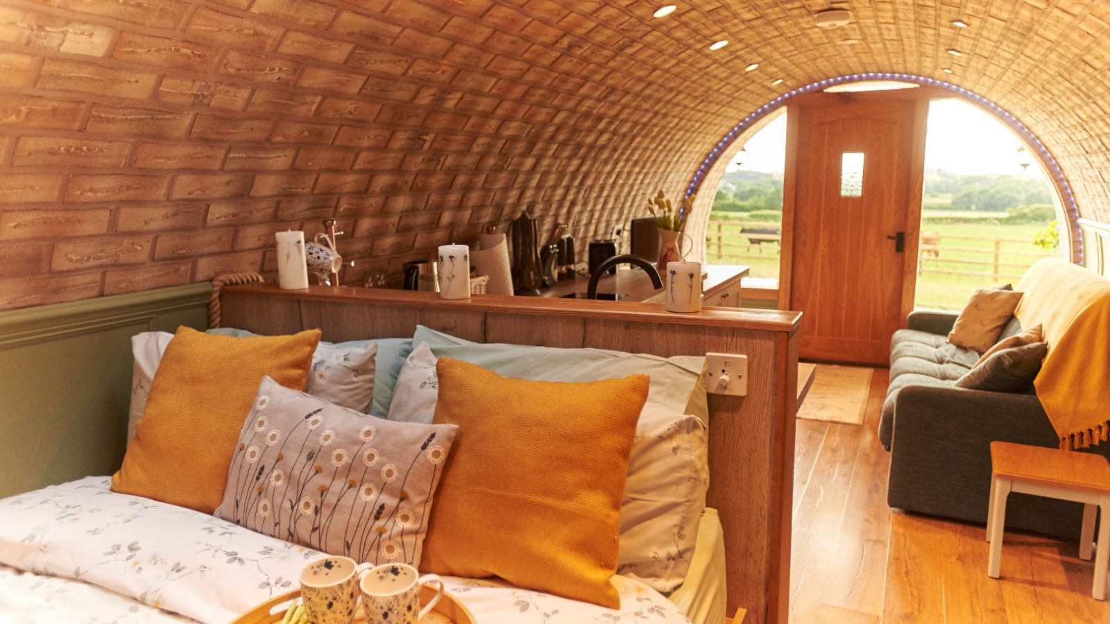 This cozy cabin interior, inspired by the charming burrows of Yorkshire, features a brick-arched ceiling, a bed adorned with yellow pillows, and a comfy sofa. Through the doorway, you'll enjoy breathtaking views of green fields that complete this idyllic retreat.