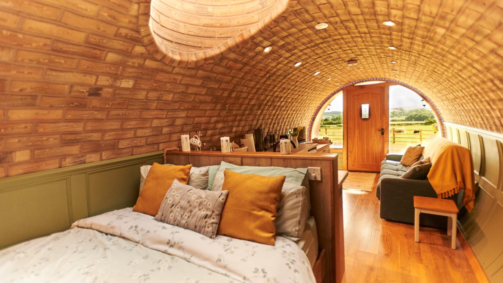 Cozy tiny home interior with a brick arched ceiling, bed, couch, and a view of the grassy Yorkshire Burrows through the front door.