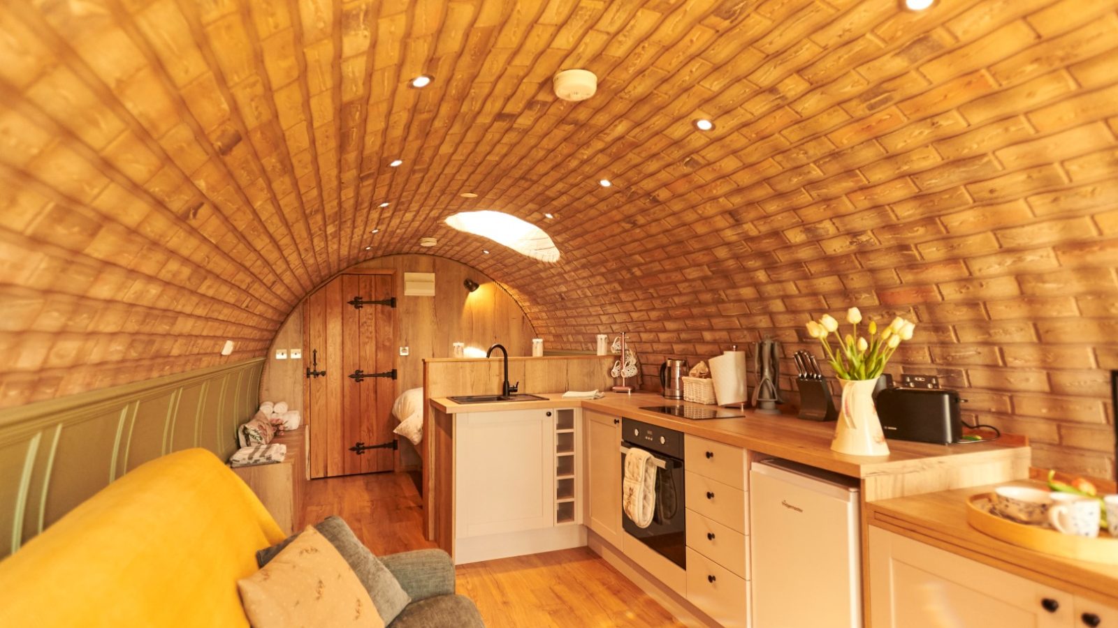 Cozy, barrel-vaulted tiny home interior reminiscent of a charming Yorkshire burrow, with wood accents. A kitchenette, sofa, and tulips on the counter glow under warm lighting that fills the space.