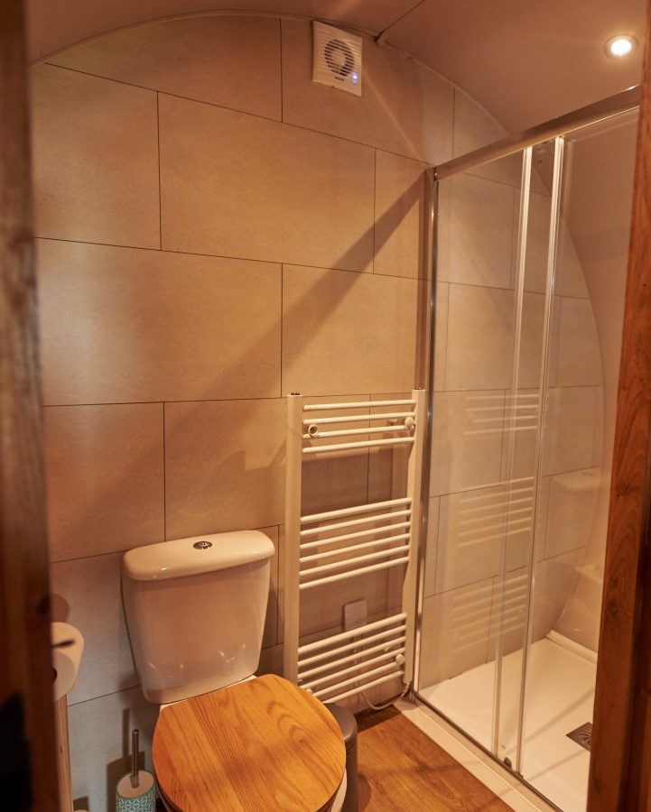 This charming bathroom features a wooden toilet seat, a convenient toilet paper holder, and a towel rack, all set against the timeless appeal of beige tiled walls. The glass-enclosed shower adds a touch of modern elegance to this quaint Yorkshire-inspired retreat.