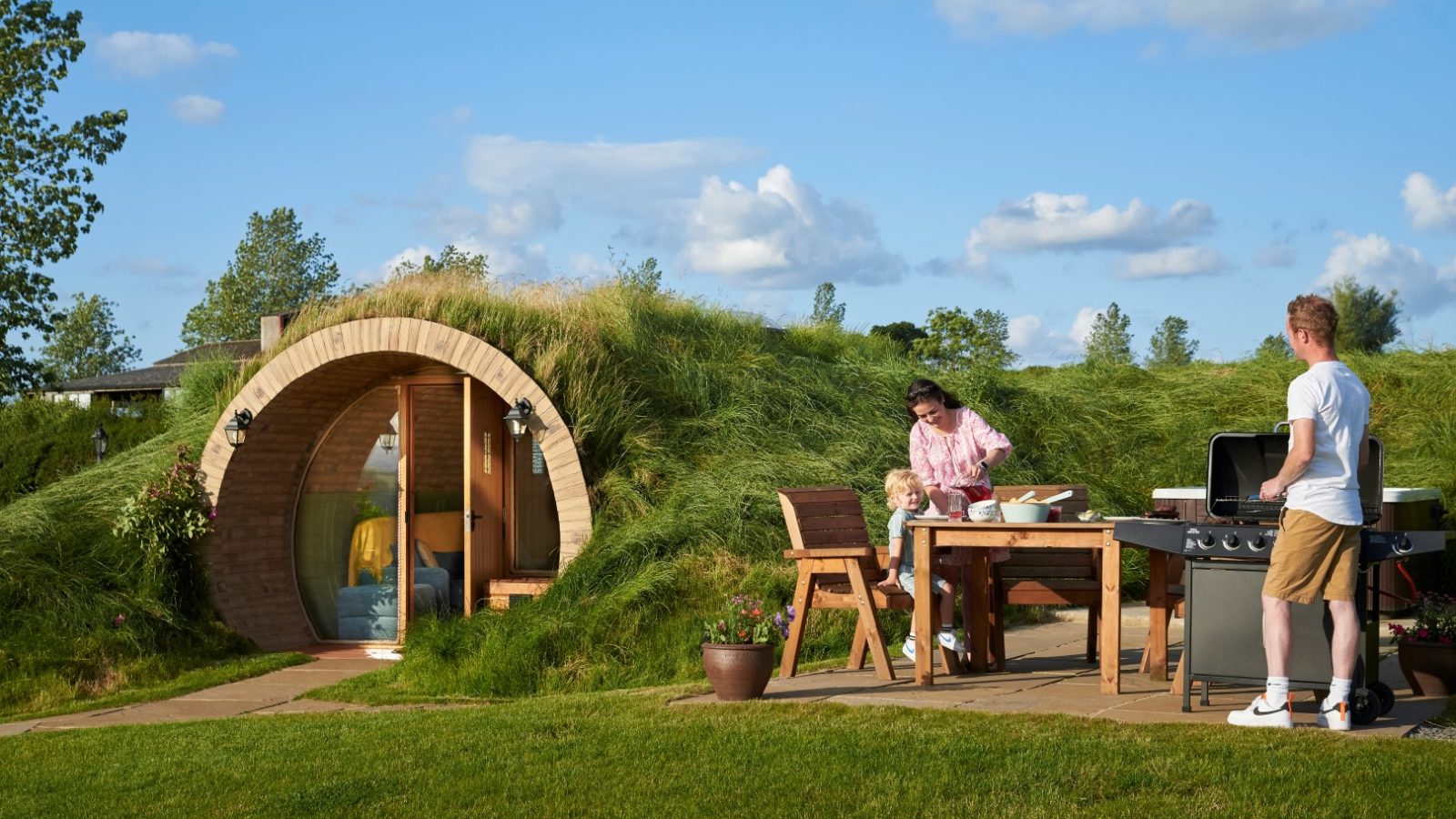 A family prepares food outside their Yorkshire Burrows-inspired, grass-covered hobbit-style home, enjoying a barbecue at the outdoor dining table.