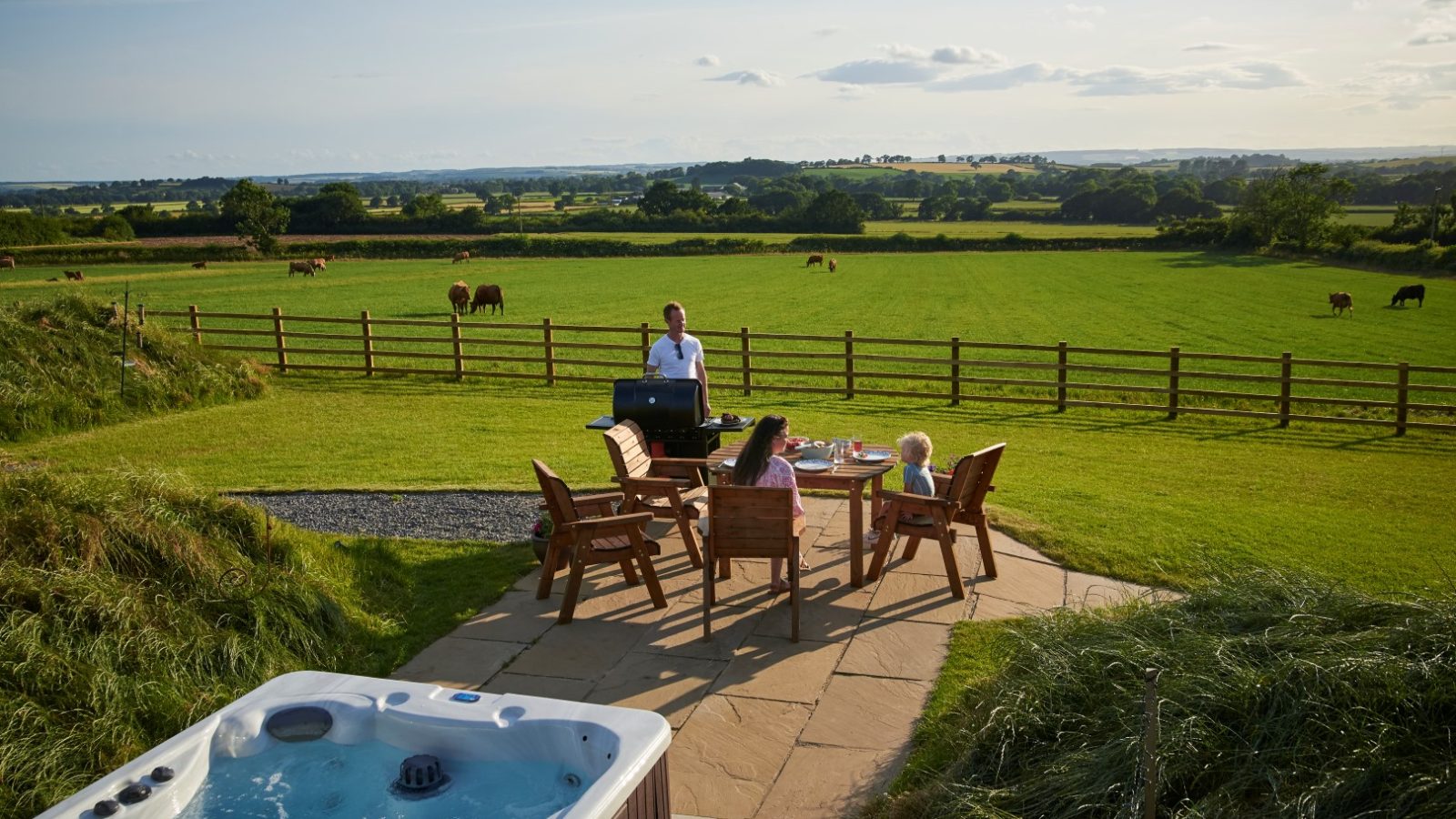 A person grills near a table with three seated people in a grassy field, reminiscent of a peaceful Yorkshire scene. A hot tub bubbles in the foreground, while cows meander leisurely in the background.