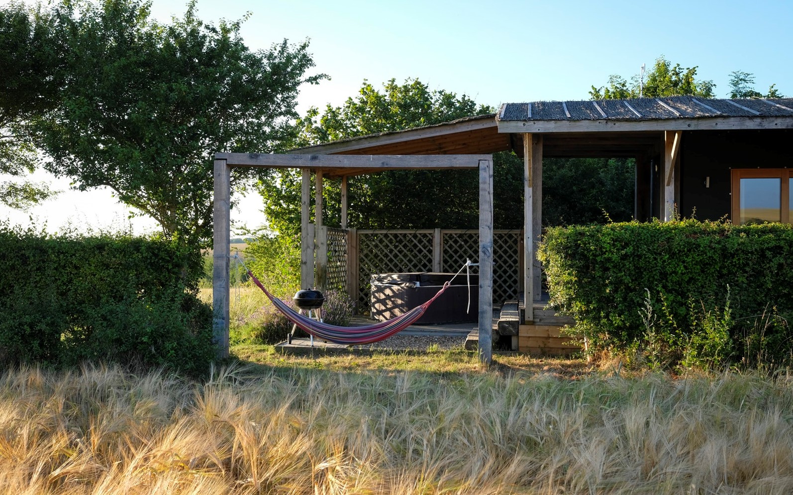 A striped hammock hangs on a wooden pergola near a rustic cabin, perfect for adults-only holidays in the UK, surrounded by lush greenery and an open field under a clear blue sky.