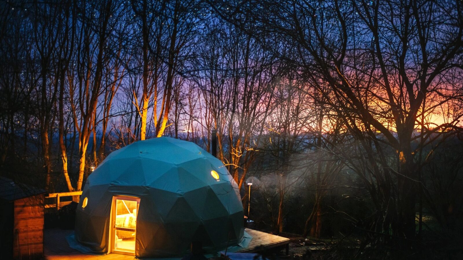 A lit geodesic dome stands in a forest clearing at night, with bare trees silhouetted against the colorful evening sky. This serene scene embodies Woodland Escapes, where the magic of Escapes Domes immerses you in nature’s tranquil beauty.