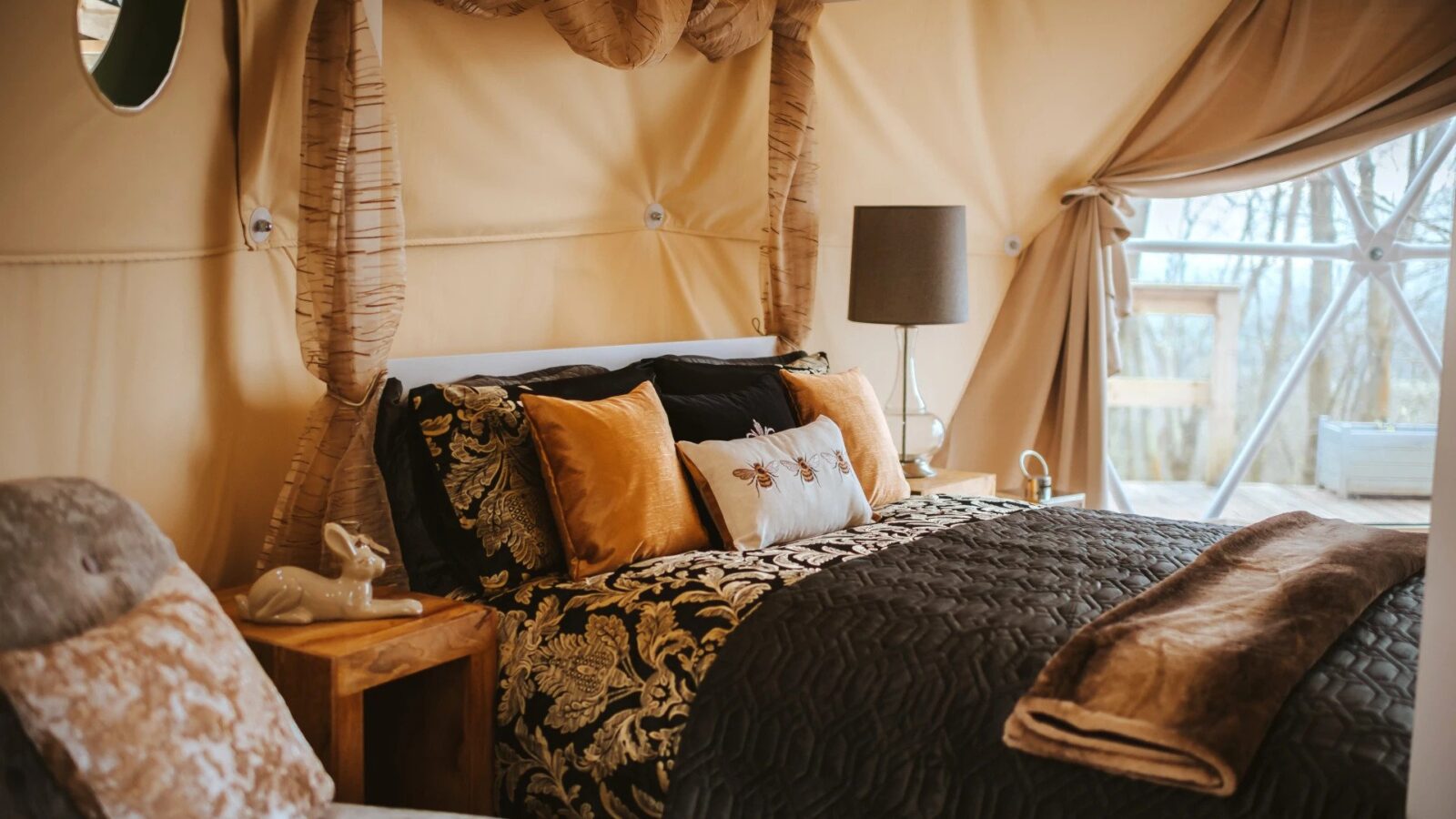 Cozy woodland glamping tent interior with a double bed, decorative pillows, a side table, and a lamp. Fabric walls add warmth to this serene escape.