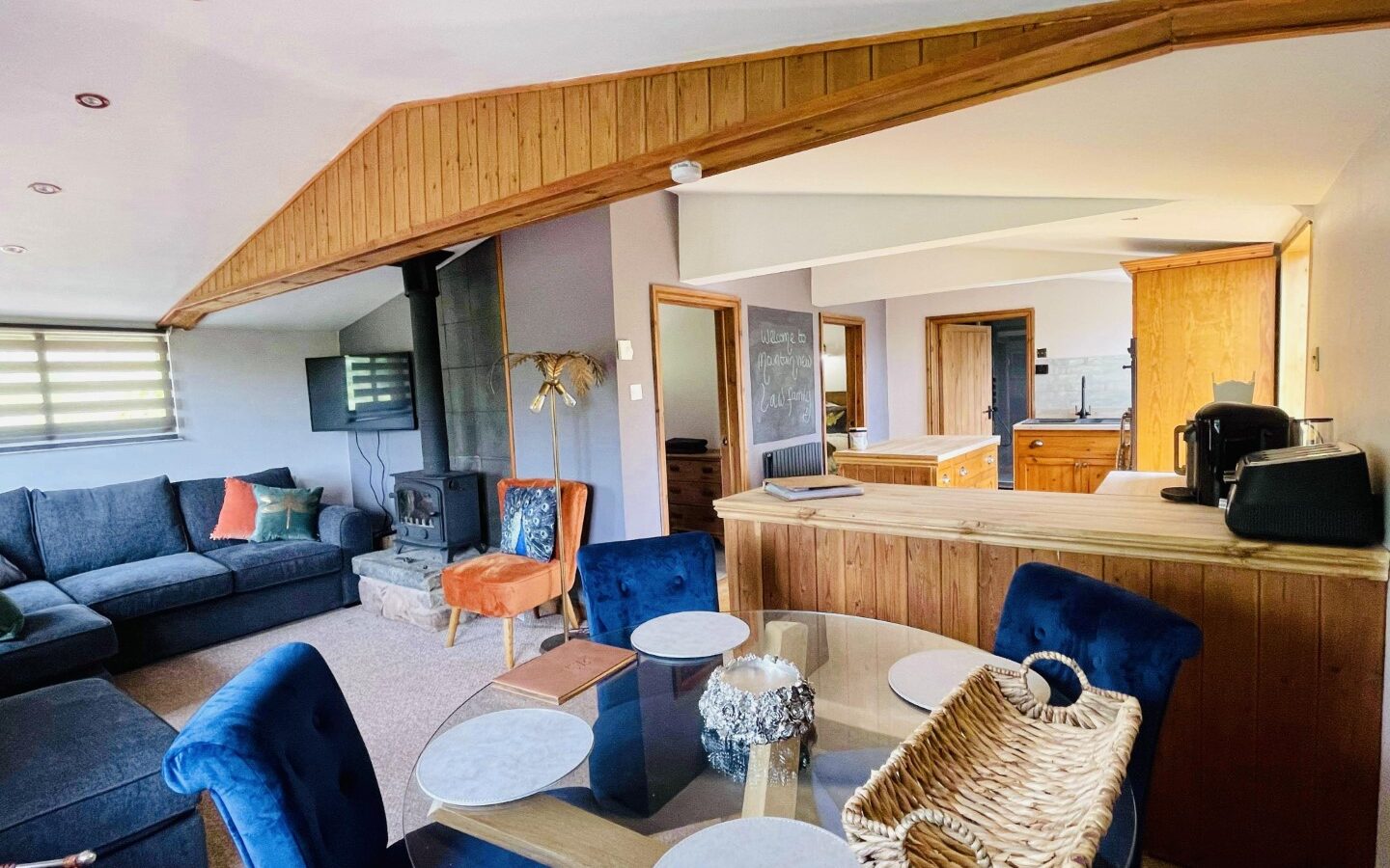 Spacious living room in this Woodland Escapes lodge, featuring blue chairs, a round glass table, gray couch, wood ceiling, and a modern kitchen nook.