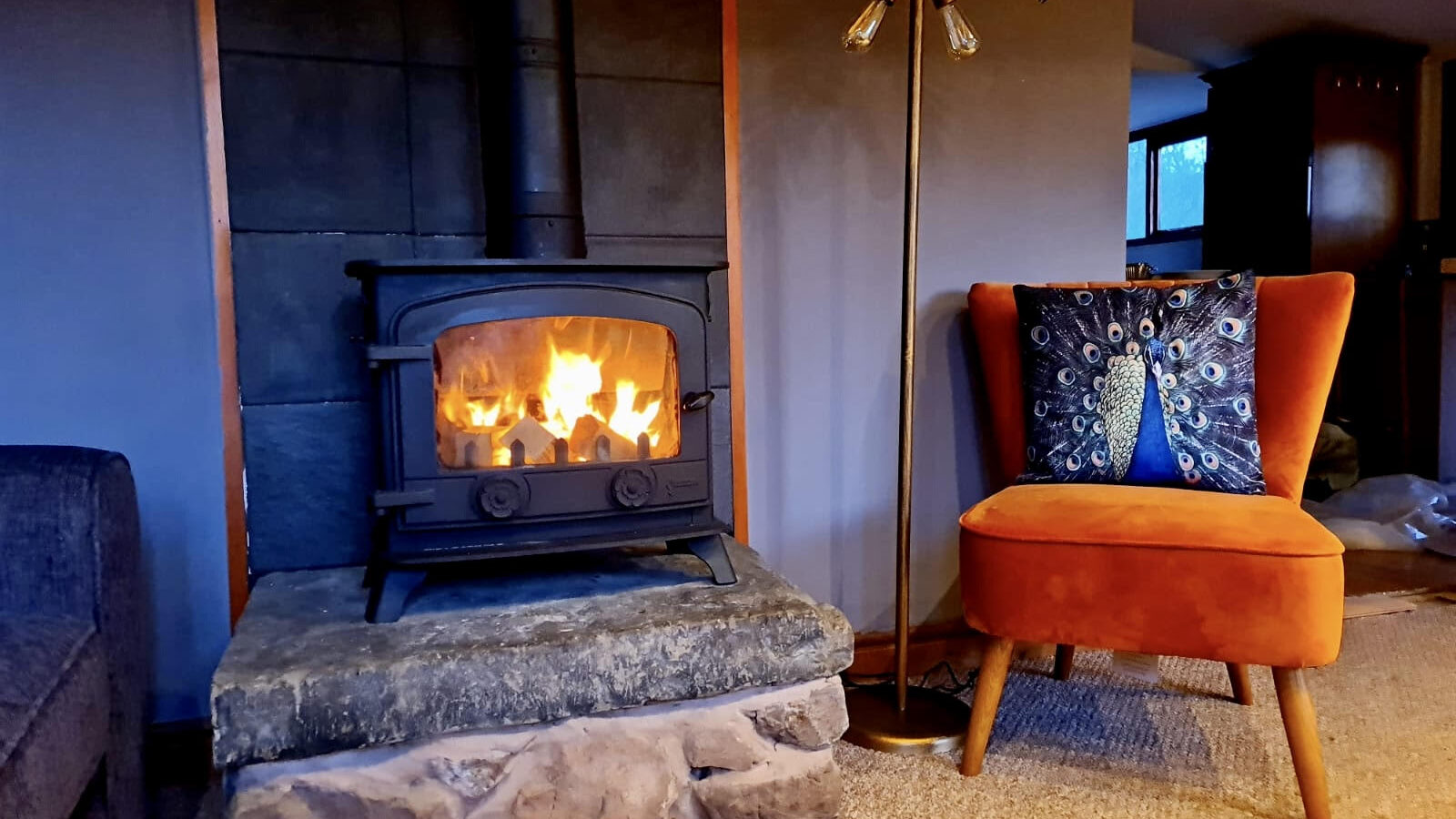 A cozy living room scene reminiscent of woodland escapes features a lit wood-burning stove, an inviting orange chair, and a decorative peacock pillow, perfect for those seeking lodge-like comfort and tranquility.