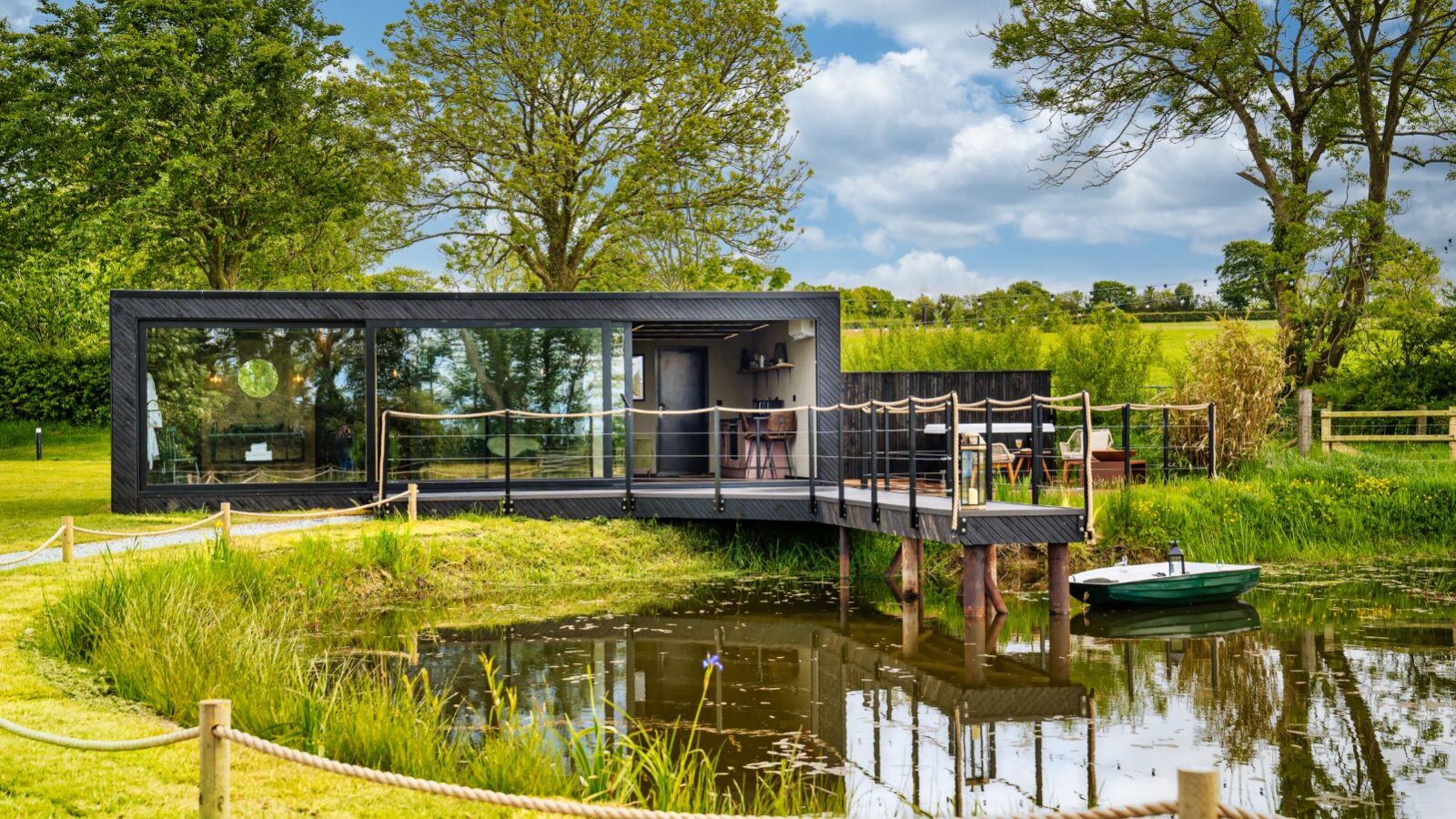 This modern house, reminiscent of a rustic boathouse, features large windows and a deck overlooking a serene pond, surrounded by lush trees and grass under a partly cloudy sky.