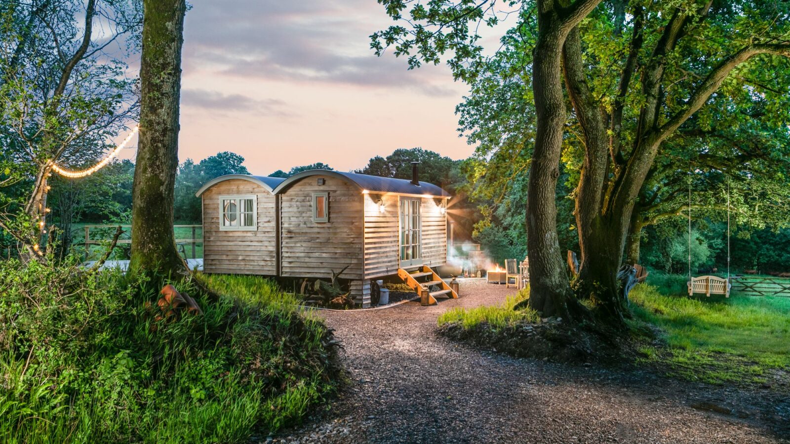 The Dimpsey offers a unique glamping experience in a wooden cabin with a curved roof, nestled among trees. Illuminated by outdoor lights, it invites you along a dirt path to your tranquil retreat.