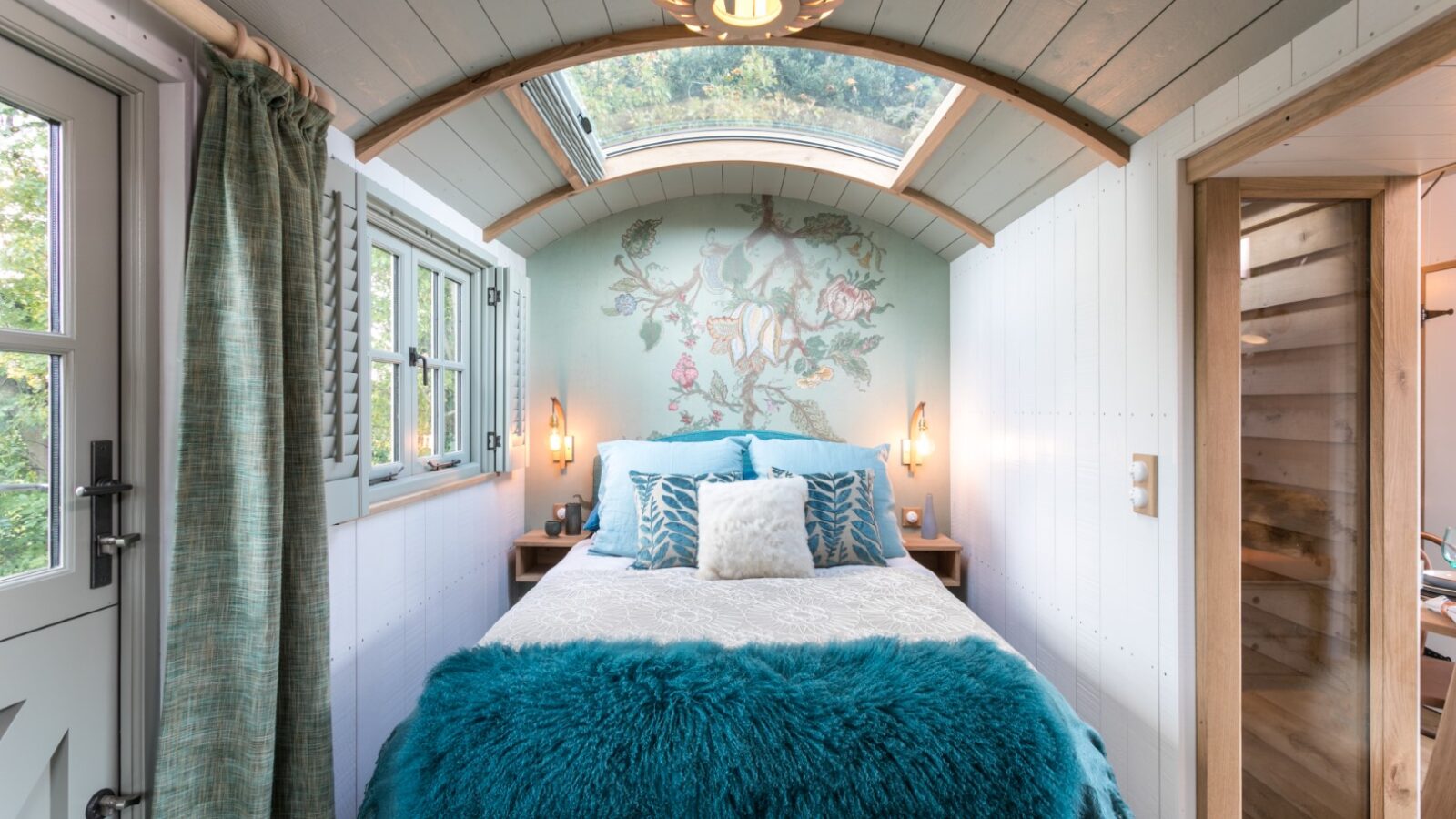 Cozy bedroom with a skylight, teal bedding, decorative pillows, and wall mural, featuring wood accents and soft lighting—perfect for a glamping vibe.