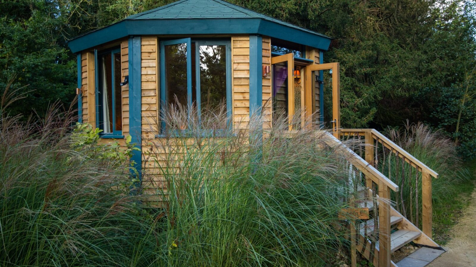 Nestled amidst tall grass and trees, this small, hexagonal wooden cabin from West Lexham Cabins features large windows and a sloped roof, offering a serene escape into nature.