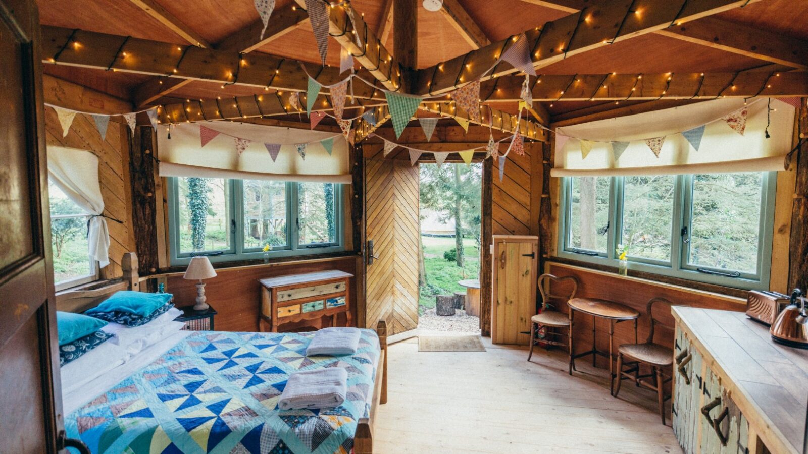 Cozy wooden cabin interior with a bed, dining table, and chairs. Decorated with bunting and string lights, reminiscent of Glamping UK style. Large windows bring the charm of West Lexham Treehouses indoors.