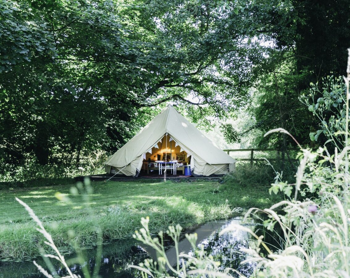 A white bell tent is set up in a lush green area near a small pond at West Lexham, with tables and chairs visible inside.