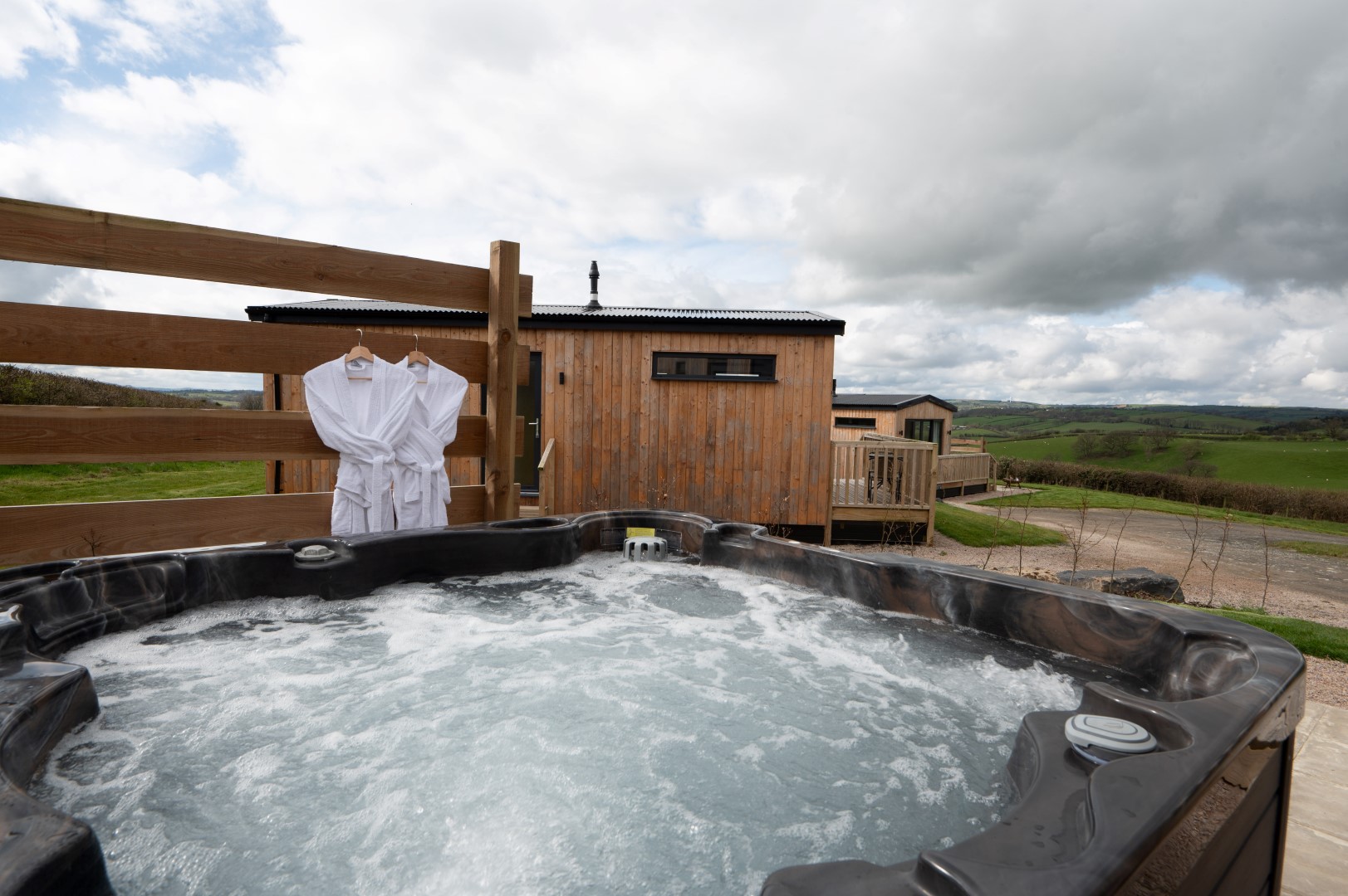 A cheap romantic retreat awaits, featuring a hot tub overlooking a scenic view with a wooden cabin nearby. Two white bathrobes hang gracefully on a wooden fence under the cloudy sky, setting the mood for an unforgettable escape.